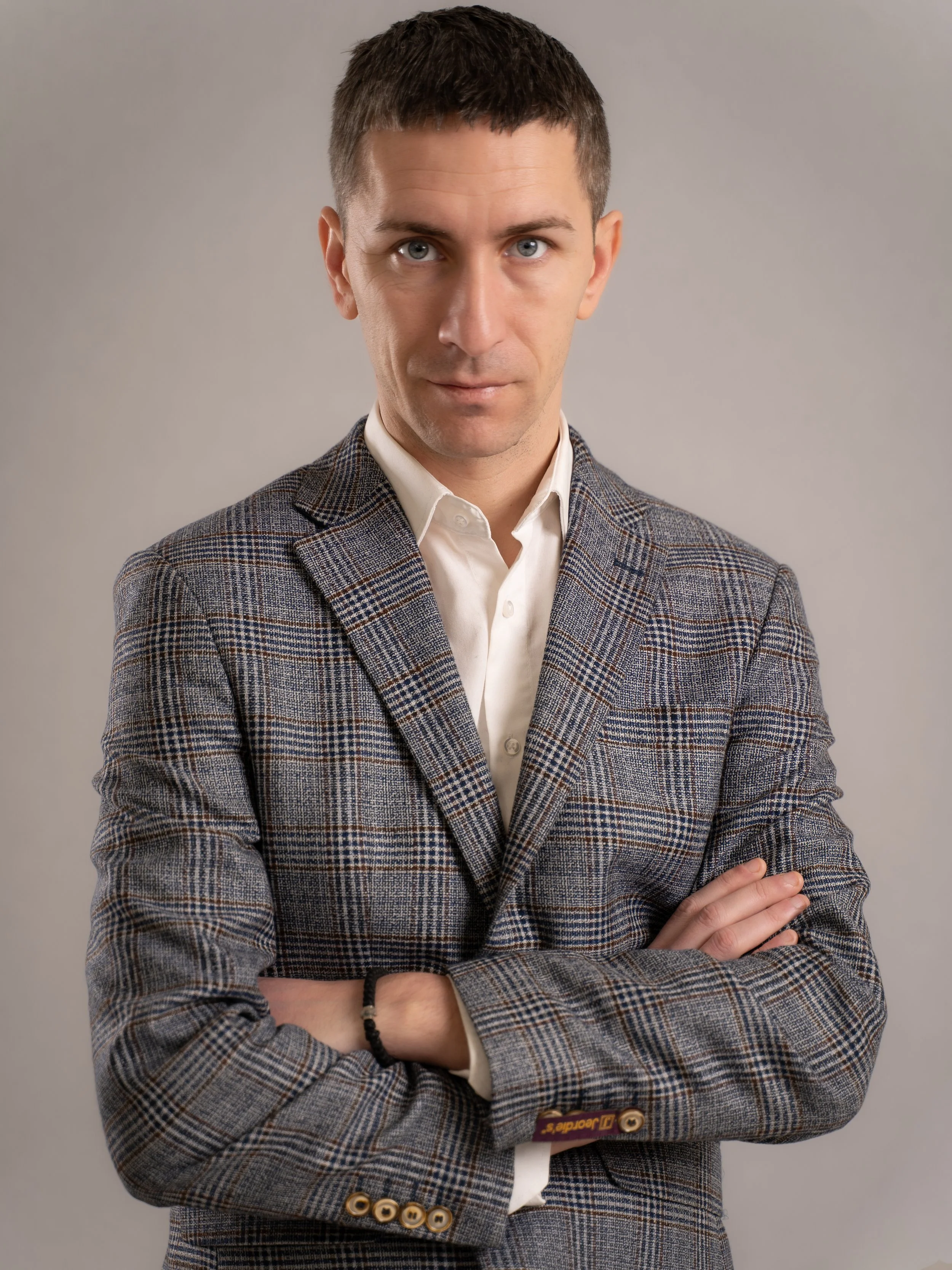 A man with short dark hair and blue eyes, wearing a checkered blazer and a white shirt, standing with arms crossed against a neutral background.