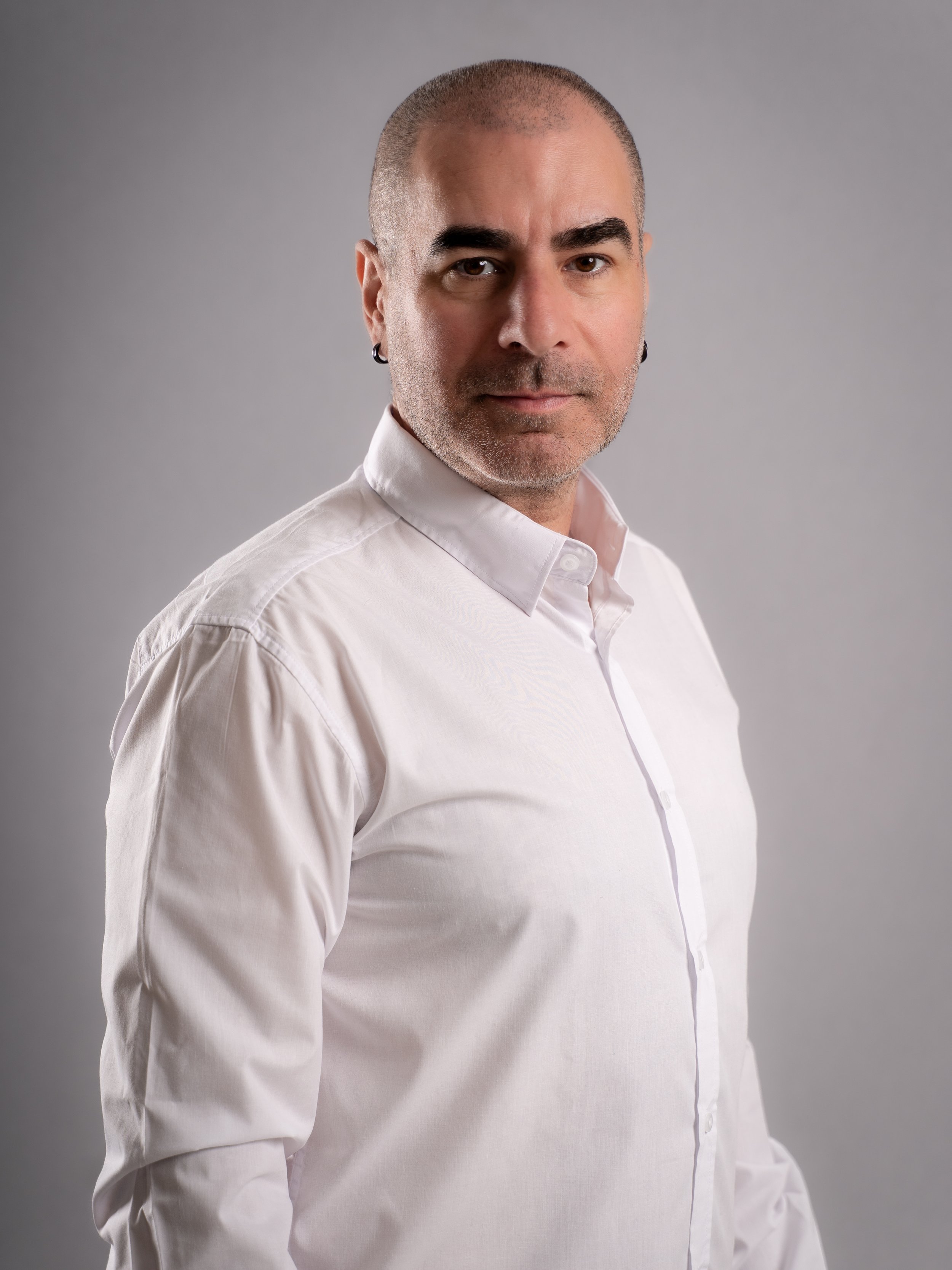 A man with a shaved head and earrings wearing a white button-up shirt, looking directly at the camera against a gray background.