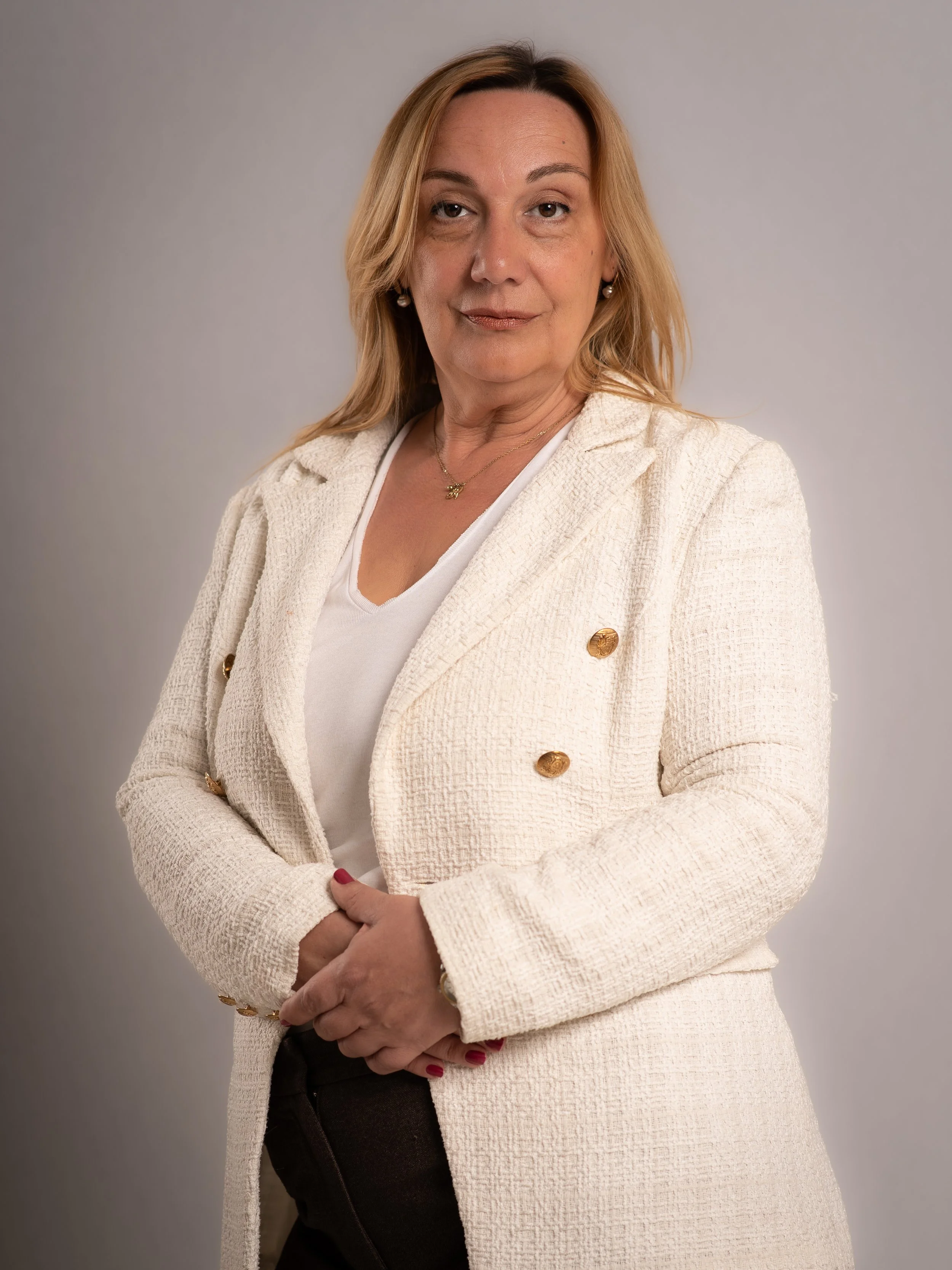 A mature woman with blonde hair wearing a cream-colored blazer with gold buttons, a white top, and dark pants, standing against a neutral background.