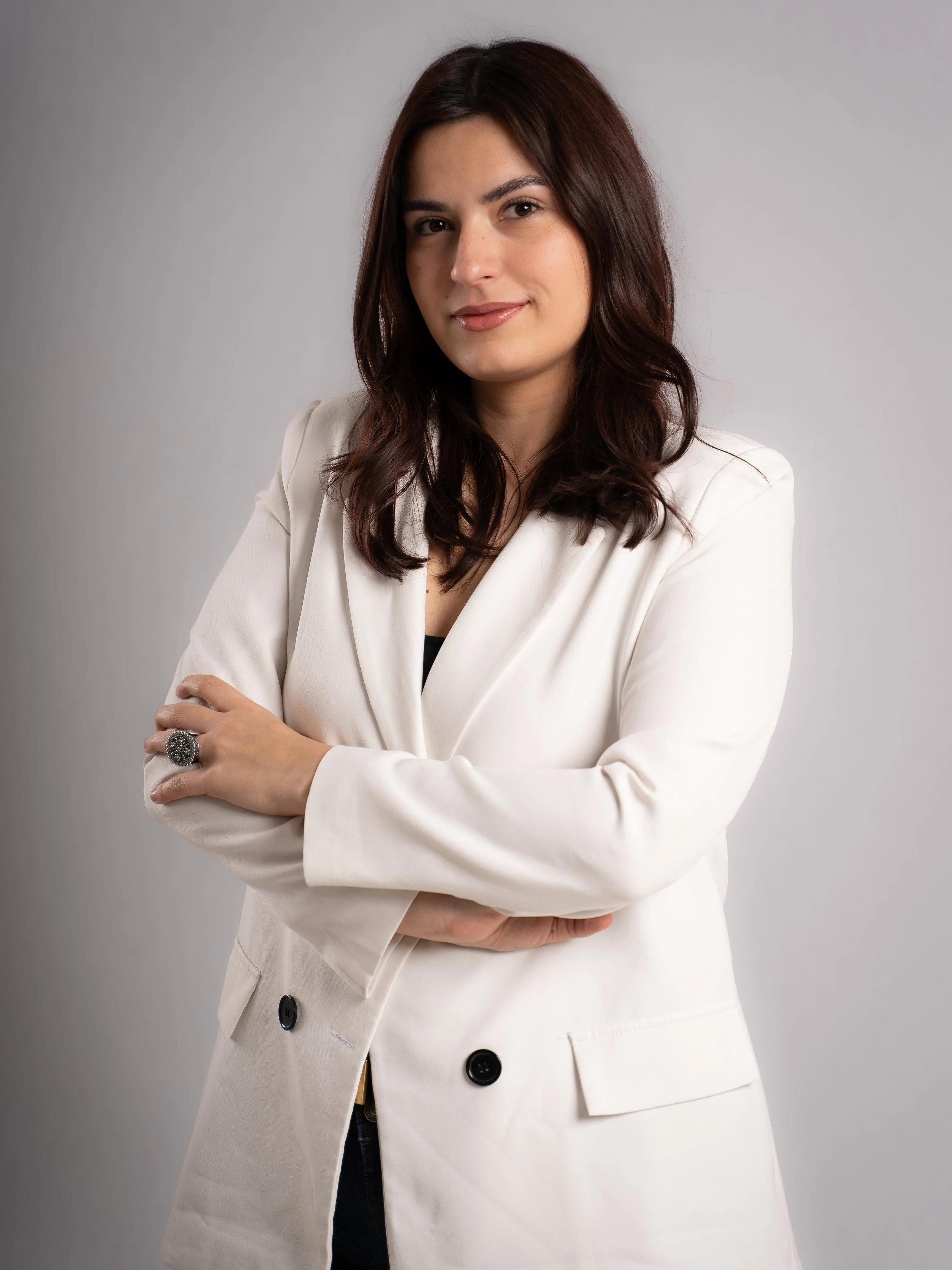 A woman with shoulder-length dark brown hair wearing a cream-colored blazer, crossing her arms, looking confident, against a plain gray background.