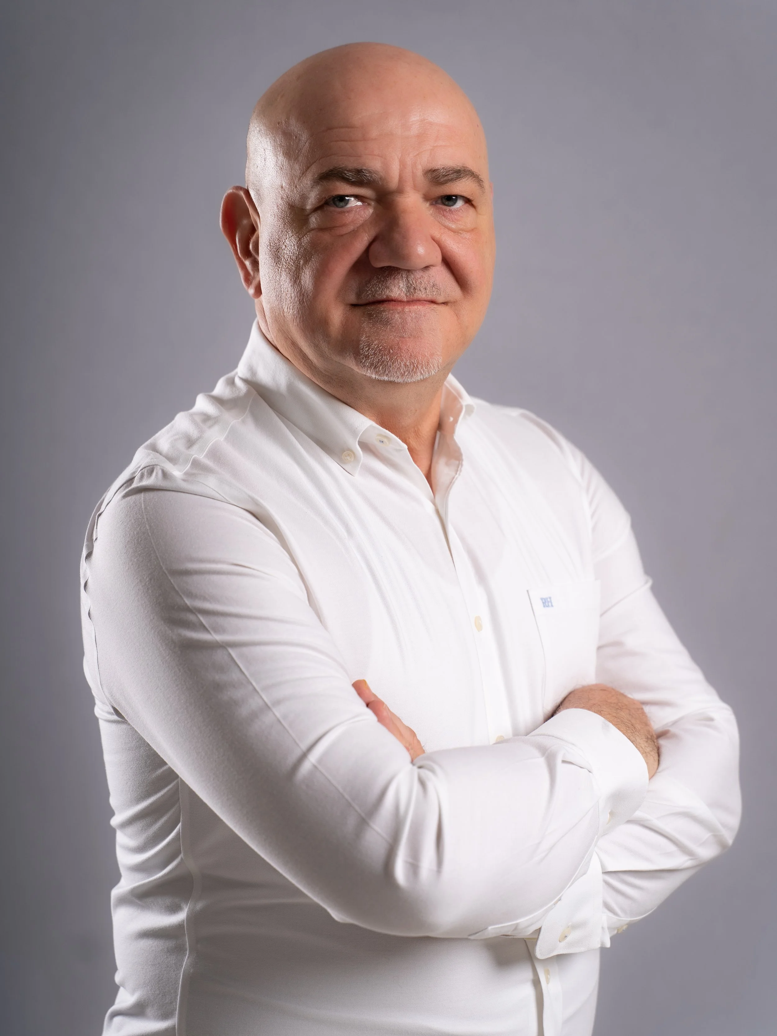 A middle-aged man with a serious expression, bald head, wearing a white dress shirt with crossed arms, standing against a plain gray background.