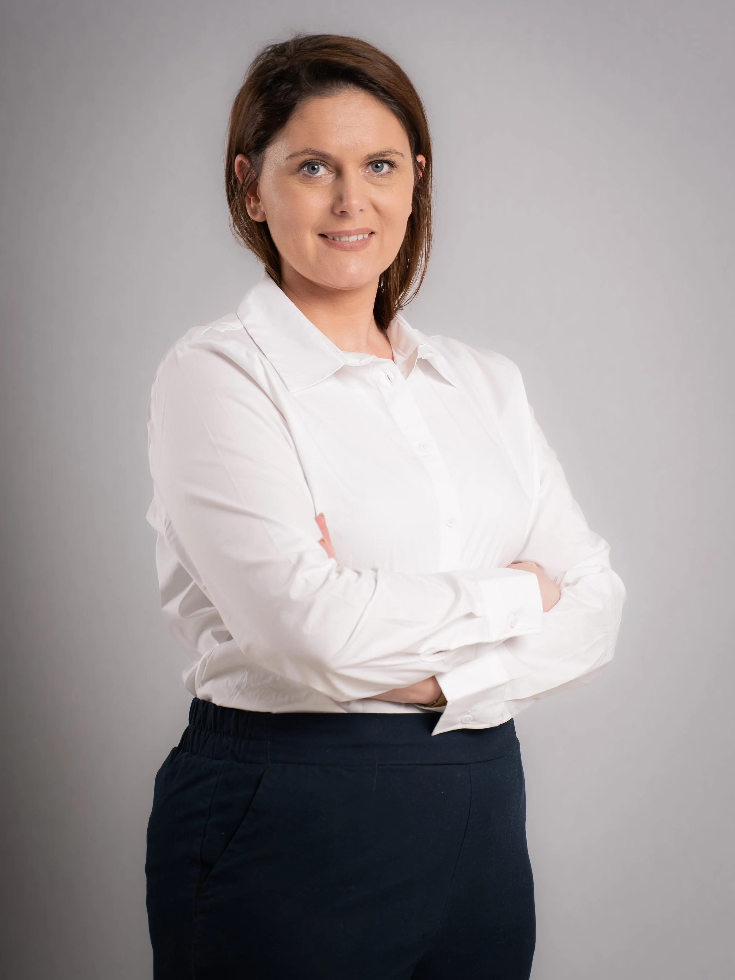 A woman with shoulder-length brown hair, wearing a white button-up shirt and dark pants, standing with arms crossed against a plain gray background.