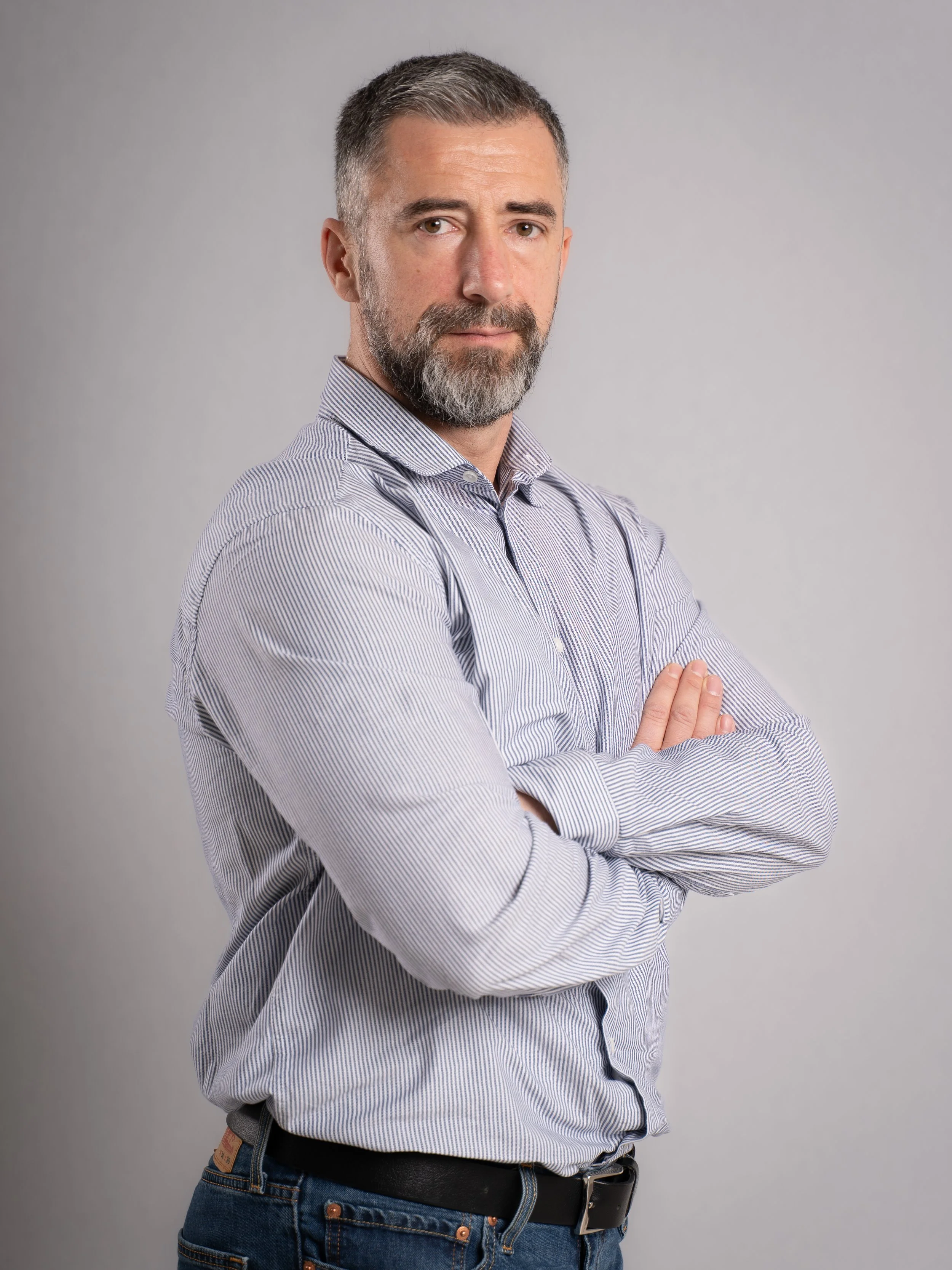A middle-aged man with gray hair and beard, wearing a striped button-up shirt and jeans, standing with arms crossed against a plain gray background.
