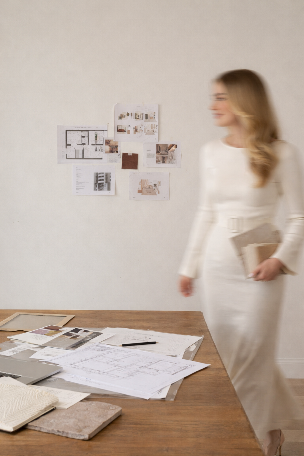 A woman in a white dress walking past a wooden table with architectural plans and sketches, with a wall of design sketches and floor plans in the background.