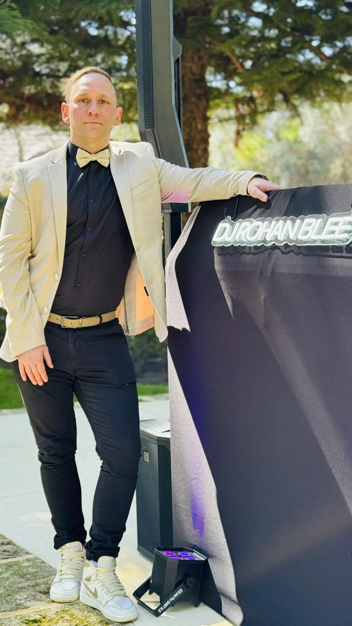 A man standing outdoors, dressed in a light-colored blazer, black shirt, beige bow tie, and sneakers, leaning on a DJ booth with a neon sign reading 'DJ RICHAN BLEE.'