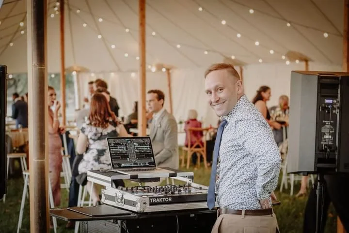 A man in a patterned shirt standing at a DJ booth with a laptop and mixer, smiling at the camera, in a decorated outdoor tent at a social gathering or wedding reception.