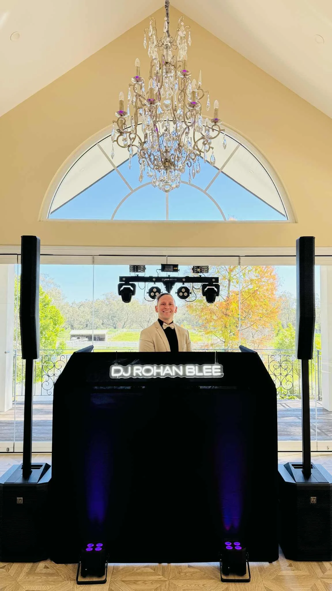 A DJ standing behind a DJ booth with a glowing sign that reads 'DJ Rohan Blee' inside a bright room with a chandelier and large arched window showing a view of trees and a blue sky.