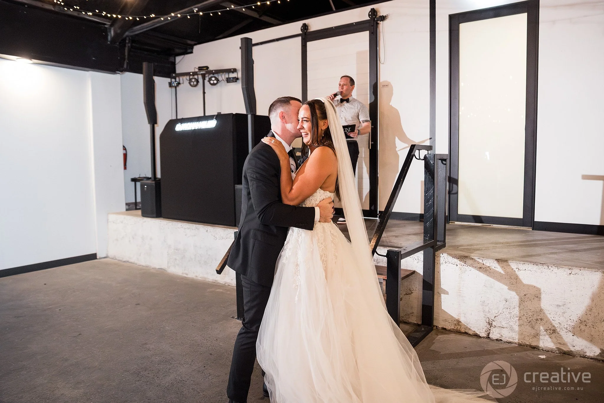 A bride and groom dancing during their wedding reception, smiling and embracing in front of a DJ booth.