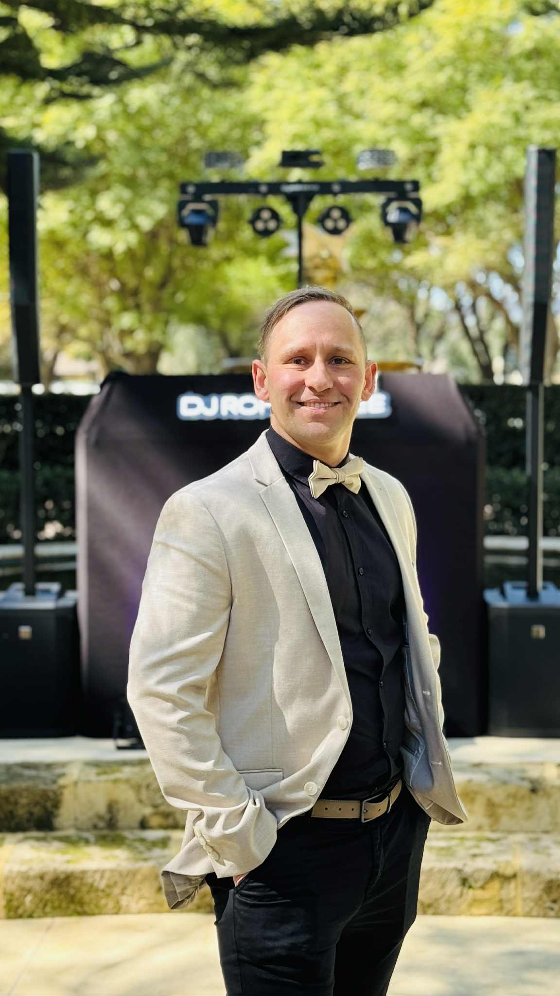 A man wearing a tuxedo jacket and a bow tie standing outdoors in front of a DJ booth at an event.