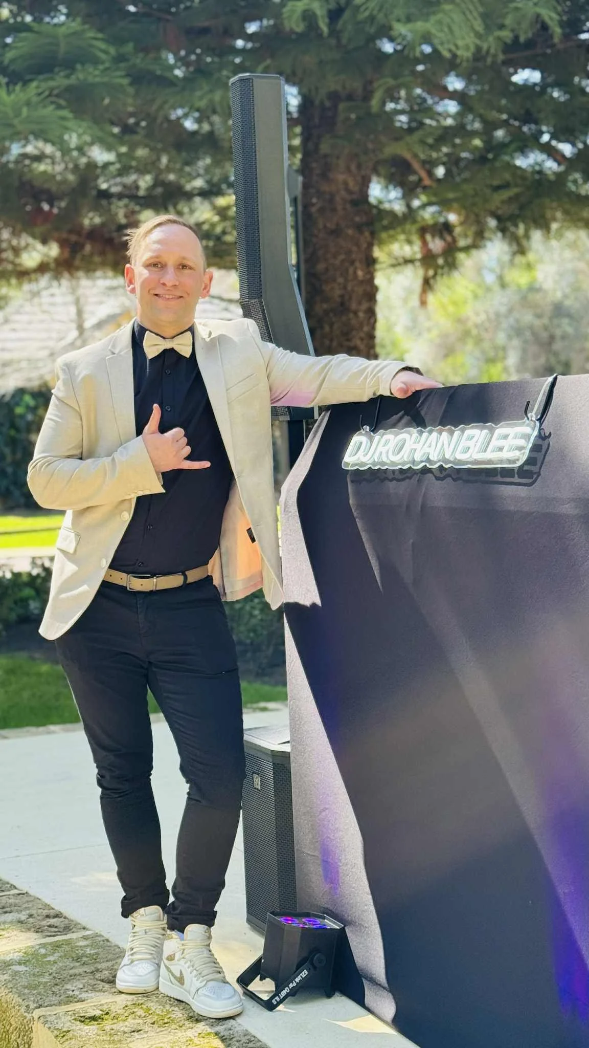 A man in a beige blazer and black shirt posing outdoors next to a black table with a illuminated sign that says 'DURCHANDLEE'. He is smiling, doing the shaka hand gesture, and has his arm around a large speaker.