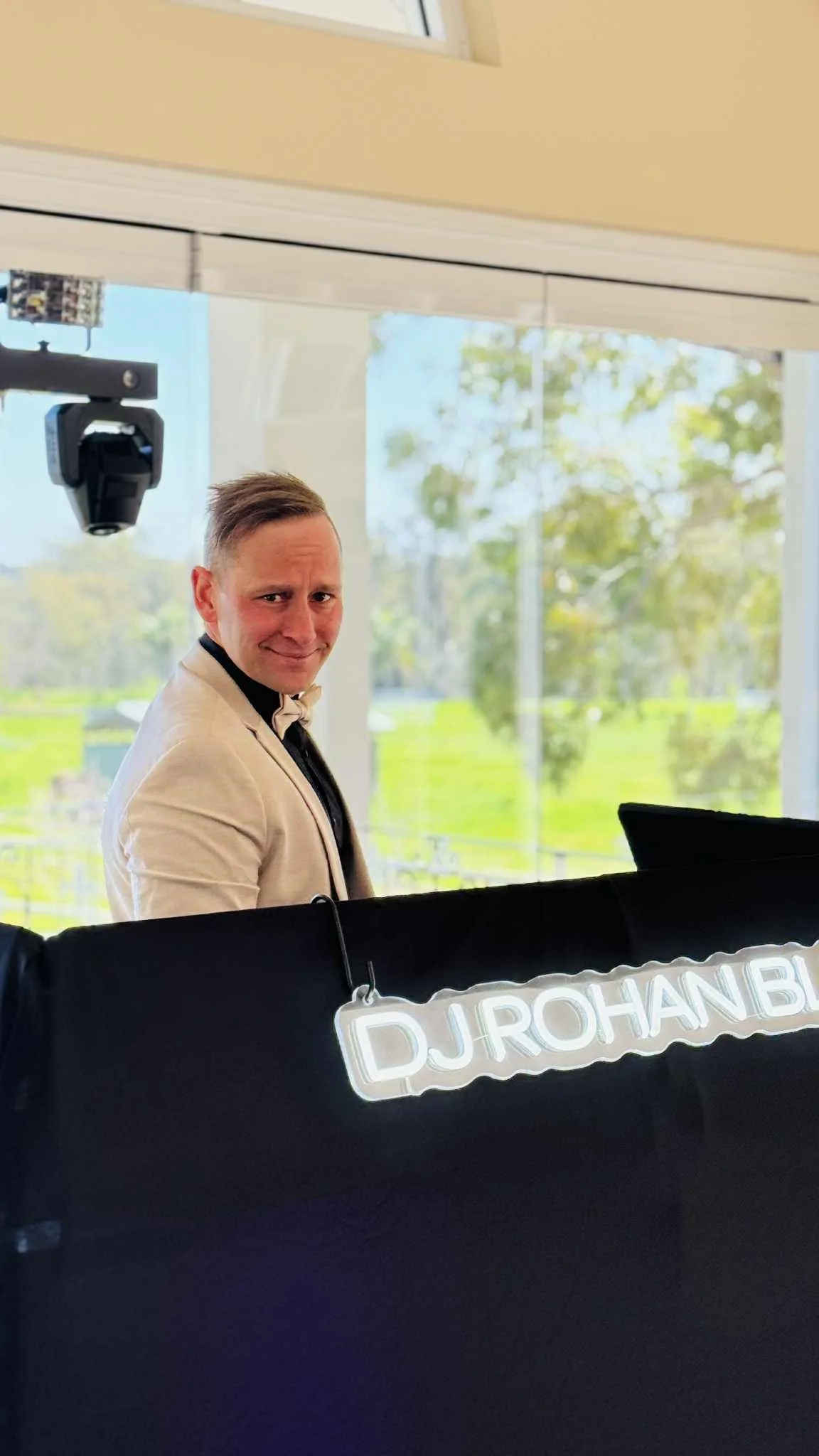 A man in a beige blazer and black shirt standing behind a black banner with a neon sign that reads 'DJ JROHAN B'. He has short, light-colored hair and is smiling, with large windows and trees in the background.