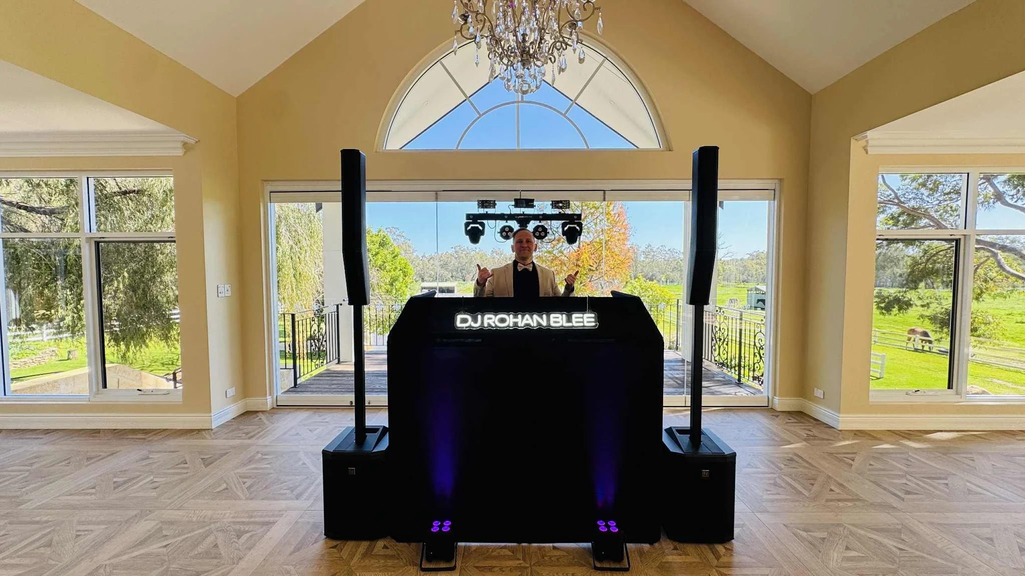 A DJ standing behind a DJ booth with a sign that says 'DJ Rohan Blee' inside a room with large windows and a view of trees and a grassy yard outside. The DJ is dressed in a tuxedo with a bow tie and is giving a thumbs-up gesture.