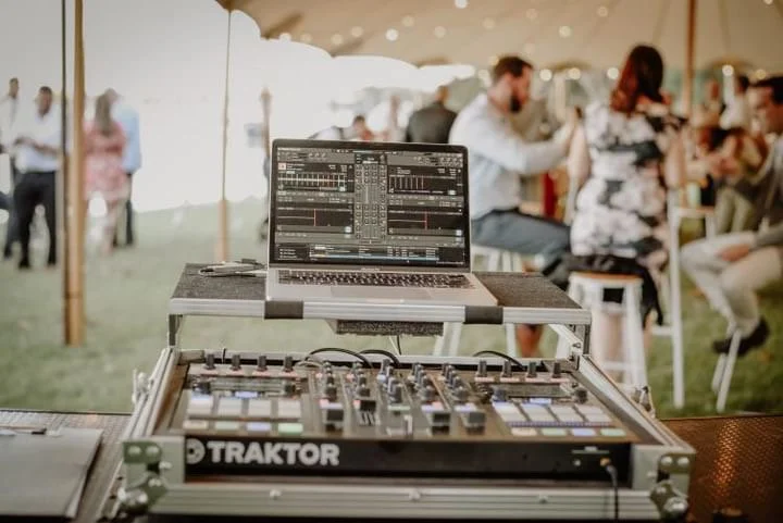 DJ equipment setup with a laptop and Traktor controller at a social event or party under a tent, with people dancing and socializing in the background.