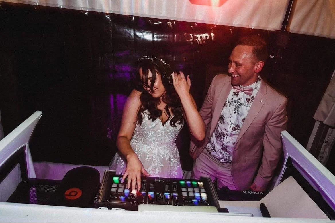 A woman in a white dress and a man in a light-colored suit and floral shirt are dancing together at a party, with the woman wearing headphones and the man smiling. They are near a DJ controller, and the woman appears to be DJing.