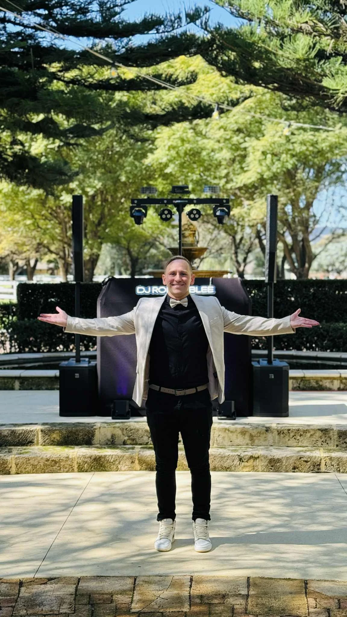 A man standing outdoors with arms outstretched in front of a DJ booth and sound equipment, surrounded by trees and sunlight.