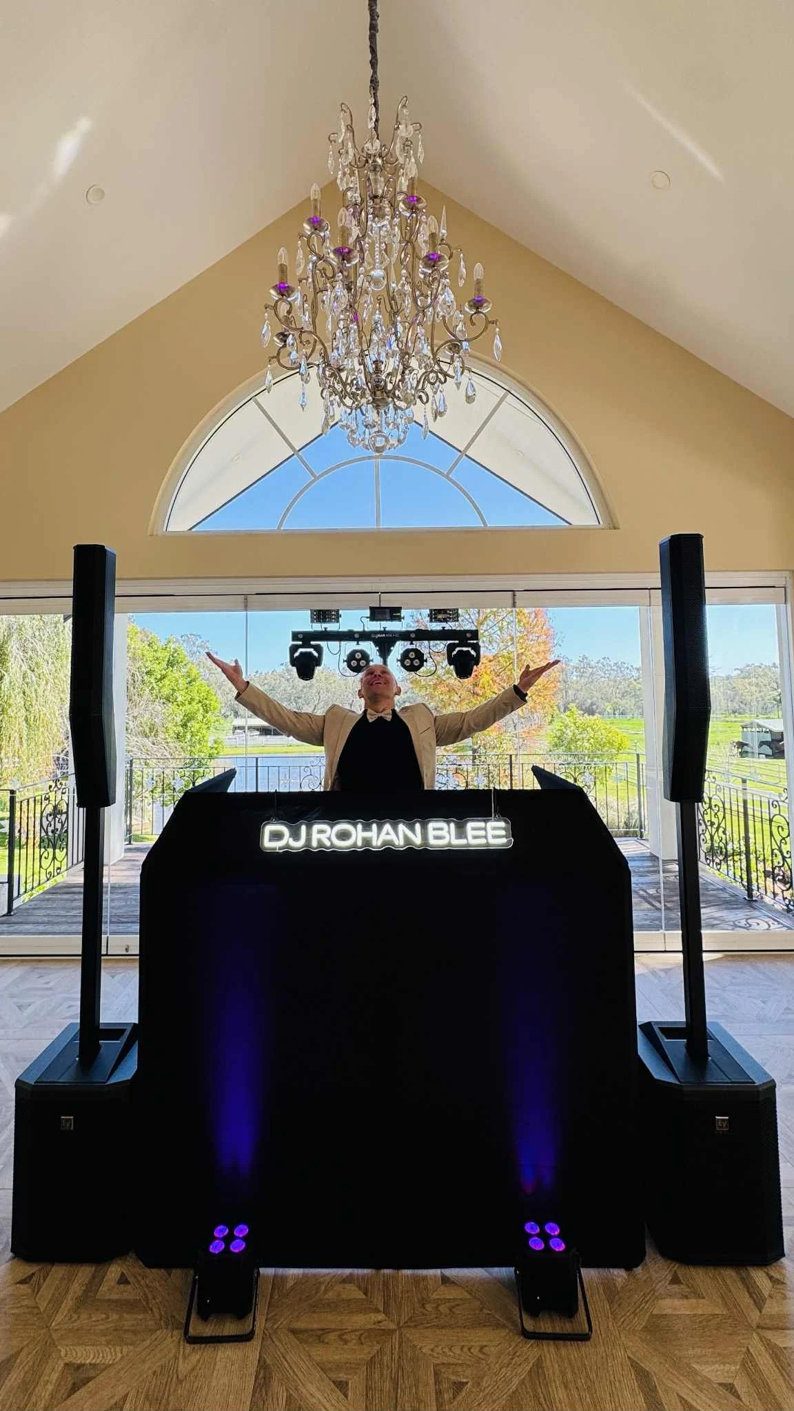 A DJ stands behind a DJ booth with a neon sign that reads 'DJ ROHAN BLEE', in a bright room with large windows, a chandelier hanging from the ceiling, and scenic outdoor views.