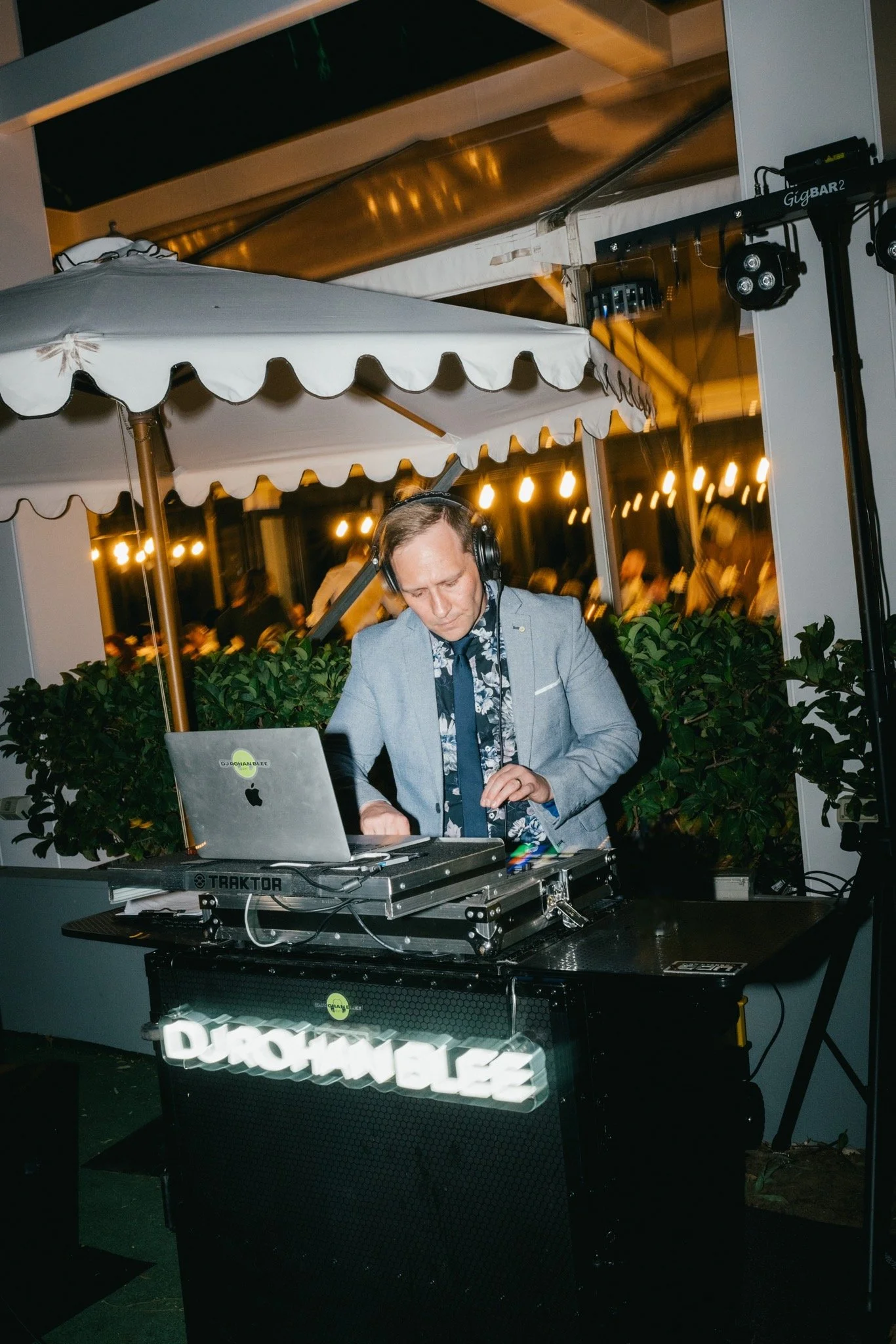 A DJ wearing a light gray blazer and headphones is mixing music on a DJ controller at an outdoor venue during the evening. There are illuminated string lights and large potted plants in the background.