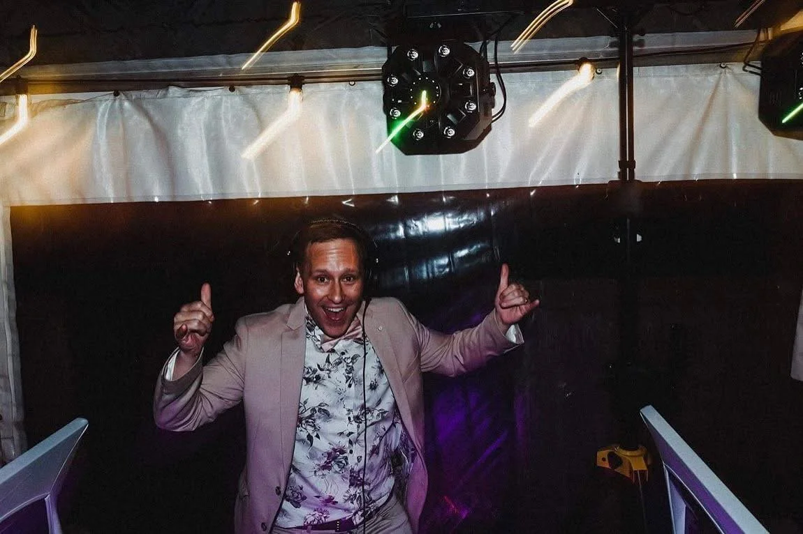 A man in a light-colored blazer and floral shirt celebrating and dancing at a party or nightclub, with his arms raised and thumbs up, under hanging ceiling lights and a black wall backdrop.