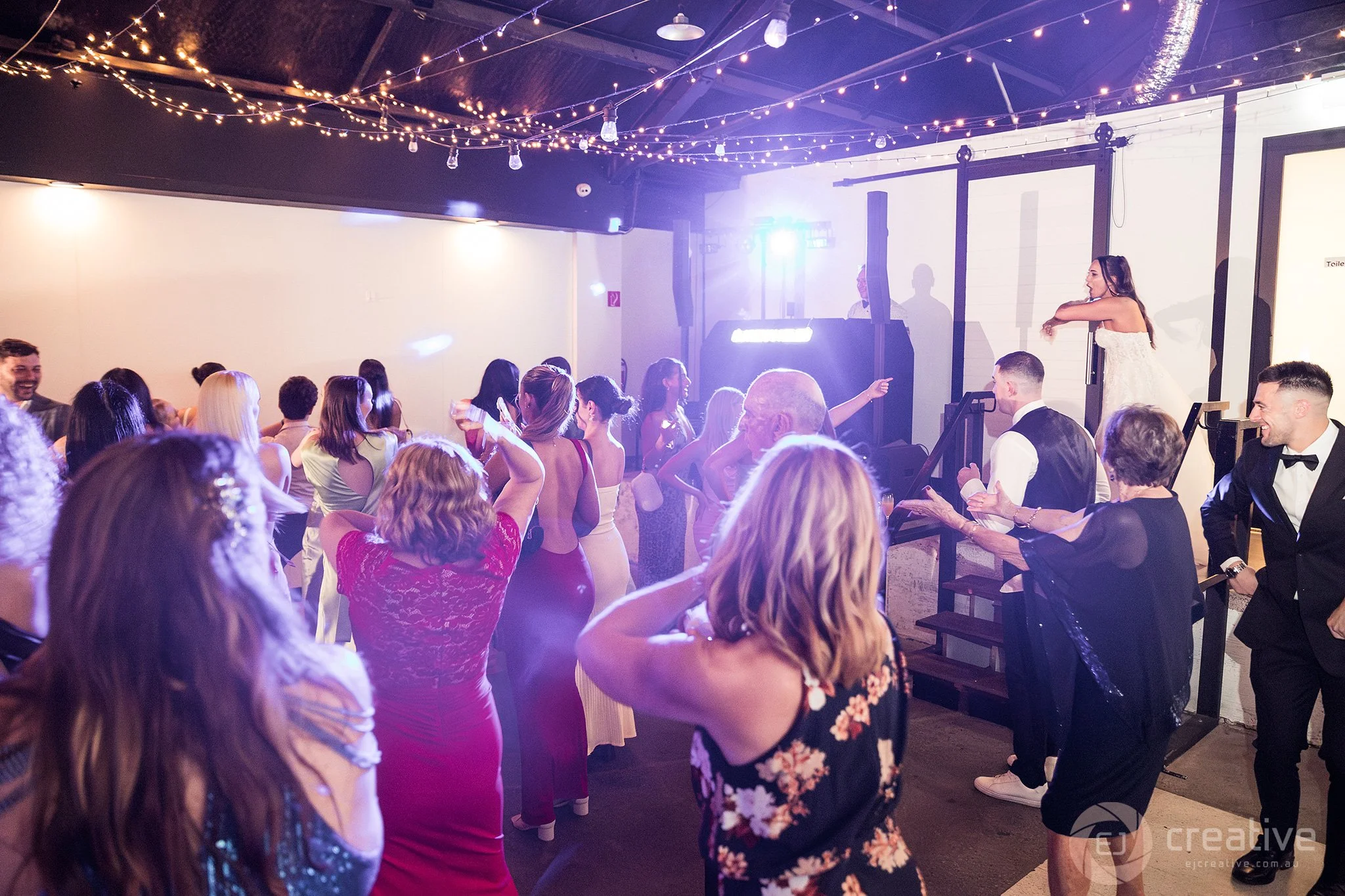 People dancing and socializing at a lively indoor event with string lights, a DJ, and a woman in a white dress dancing on a raised platform.