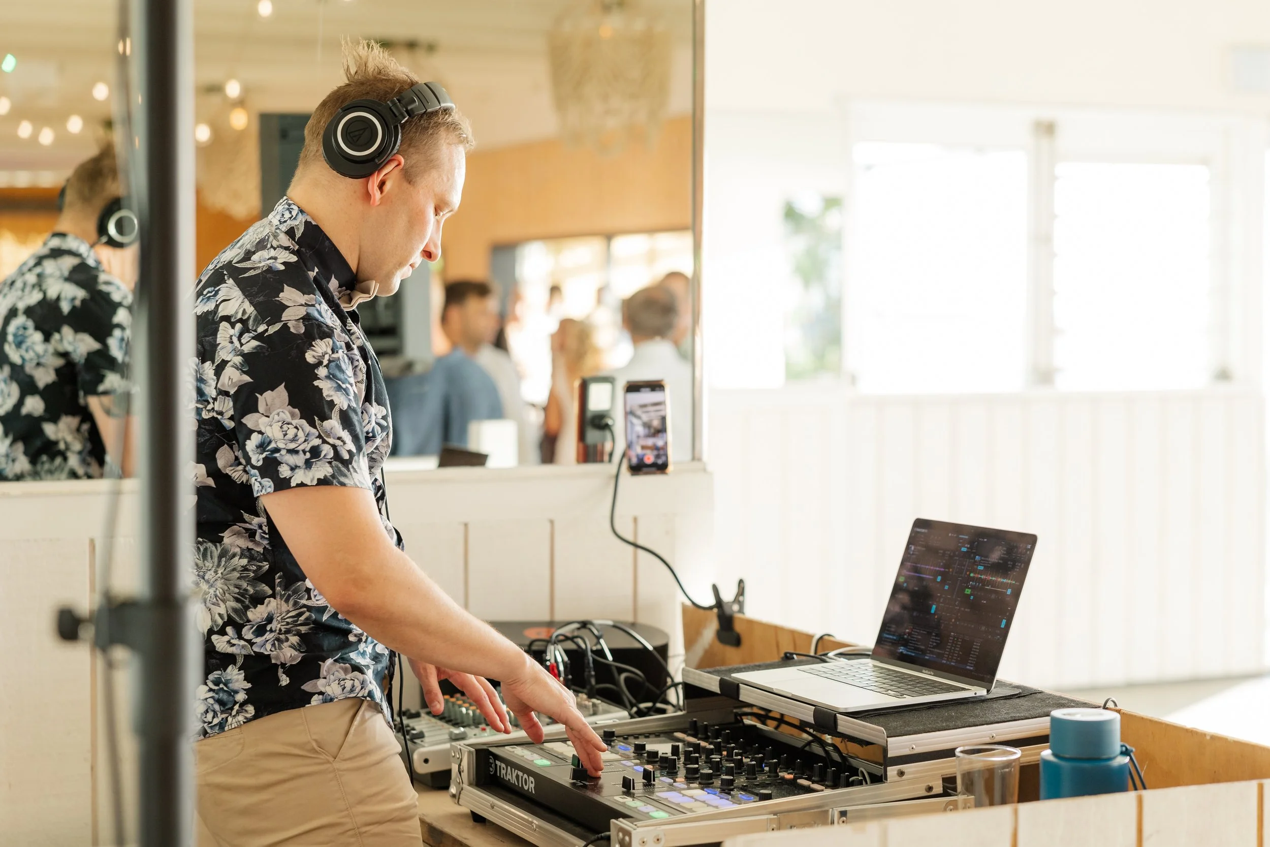 A young man wearing headphones and a floral shirt DJing at an event, with a laptop, mixing equipment, and a smartphone mounted on a stand in front of him. People are mingling in the background.