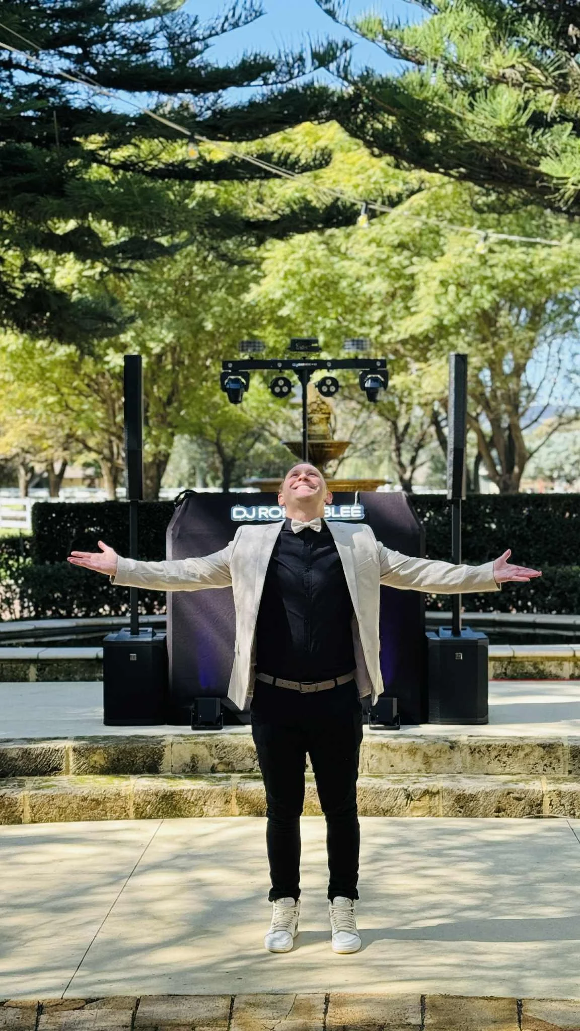 A person stands outdoors with arms wide open, smiling and looking upward in front of DJ equipment set up on a stage, surrounded by trees and clear sky.