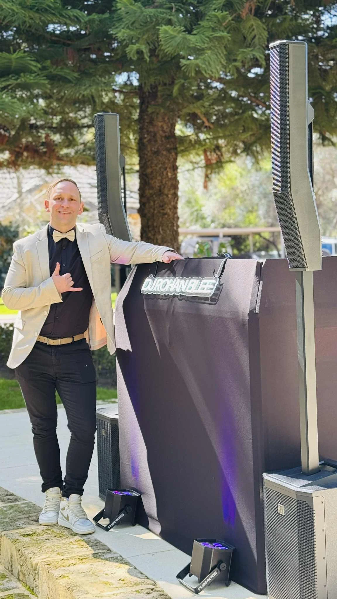 A man in a tuxedo and beige blazer standing next to DJ equipment outdoors, with trees in the background.