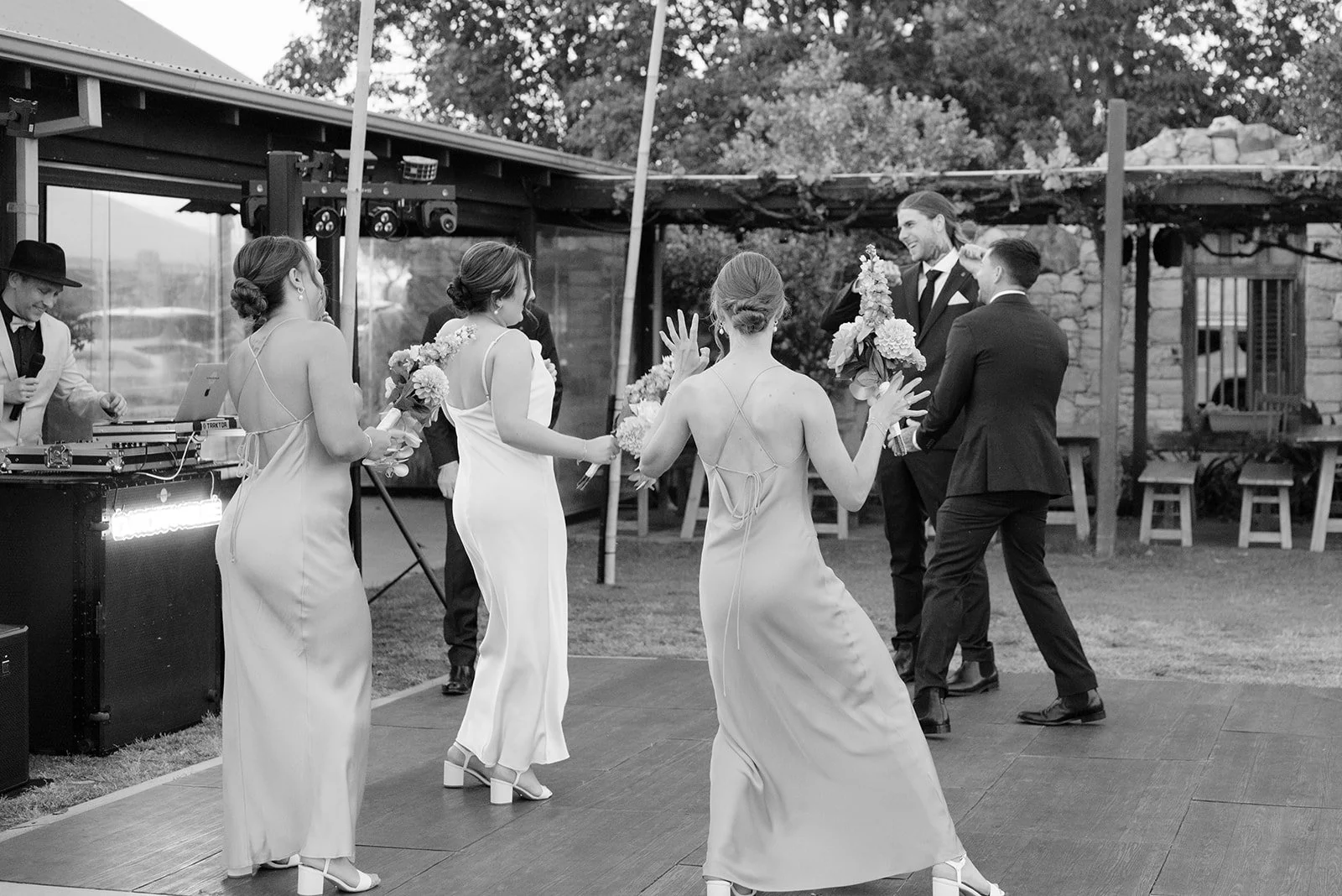 Black and white photo of a wedding reception dance floor with bridesmaids in light-colored dresses and groomsmen in suits, dancing and holding bouquets, in an outdoor venue with a DJ and a stone wall in the background.