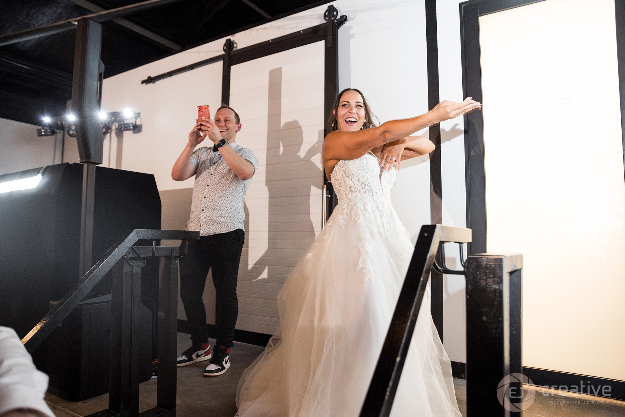 A bride in a white wedding gown smiling and dancing with her arms extended, with a man in a short-sleeved shirt taking a photo or video of her on a smartphone, in a venue with black and white decor.