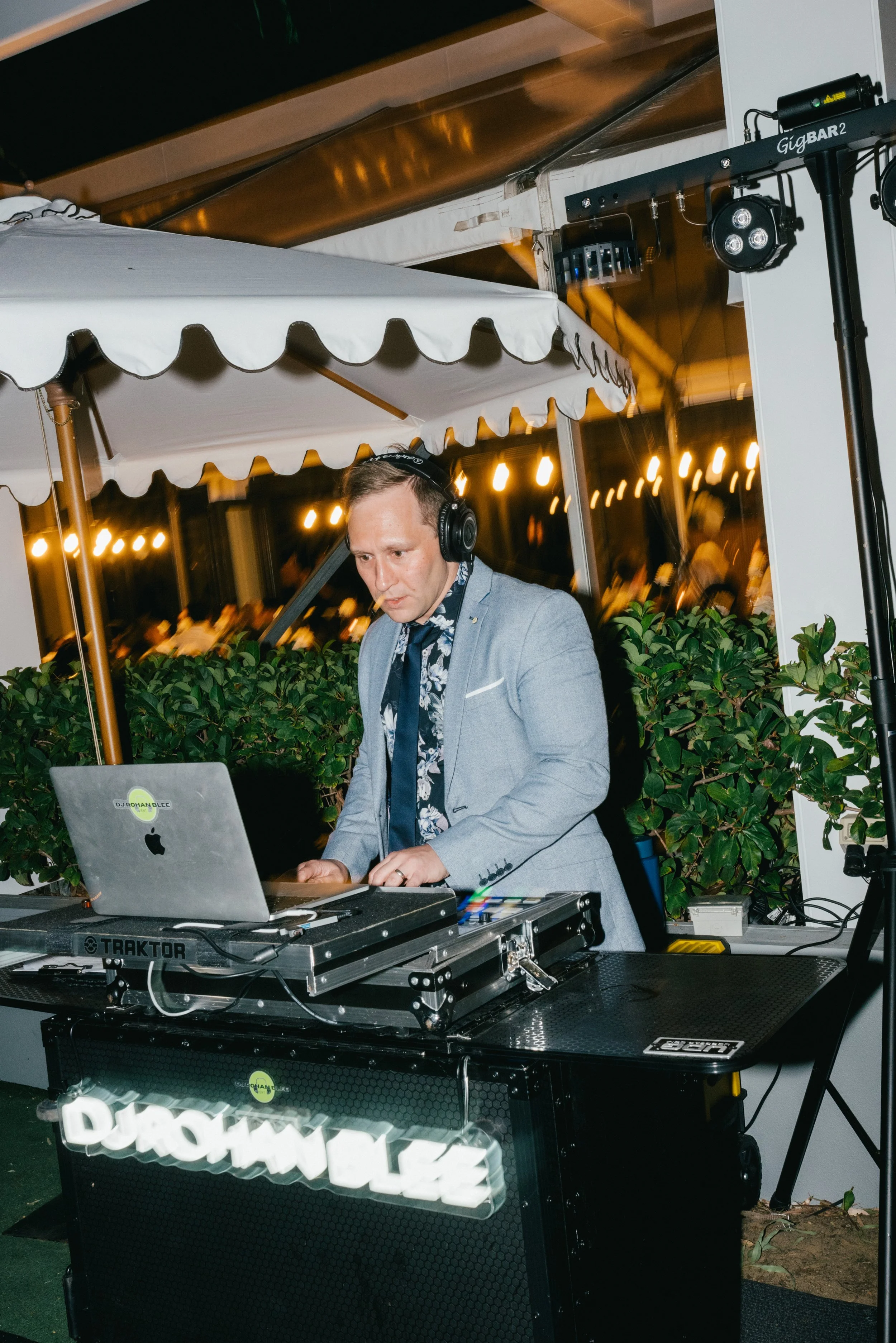 A DJ wearing a grey blazer, floral shirt, and headphones, performing at an outdoor event during the evening, with lights and trees in the background.