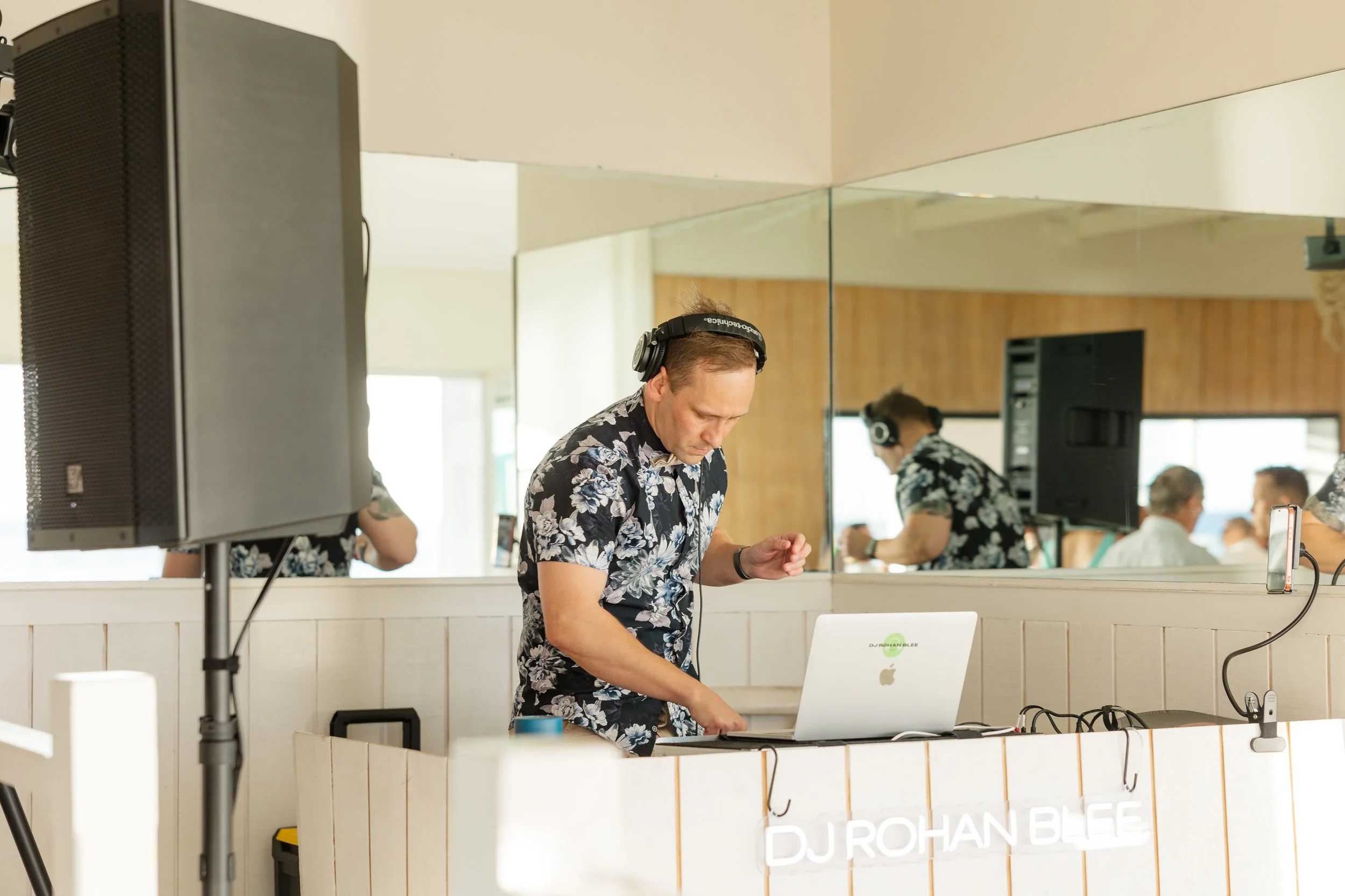 DJ Rohan Blee wearing a floral shirt and headphones, mixing music at a DJ booth with a laptop, in a room with a mirror wall and wood paneling, music equipment, and people in the background.