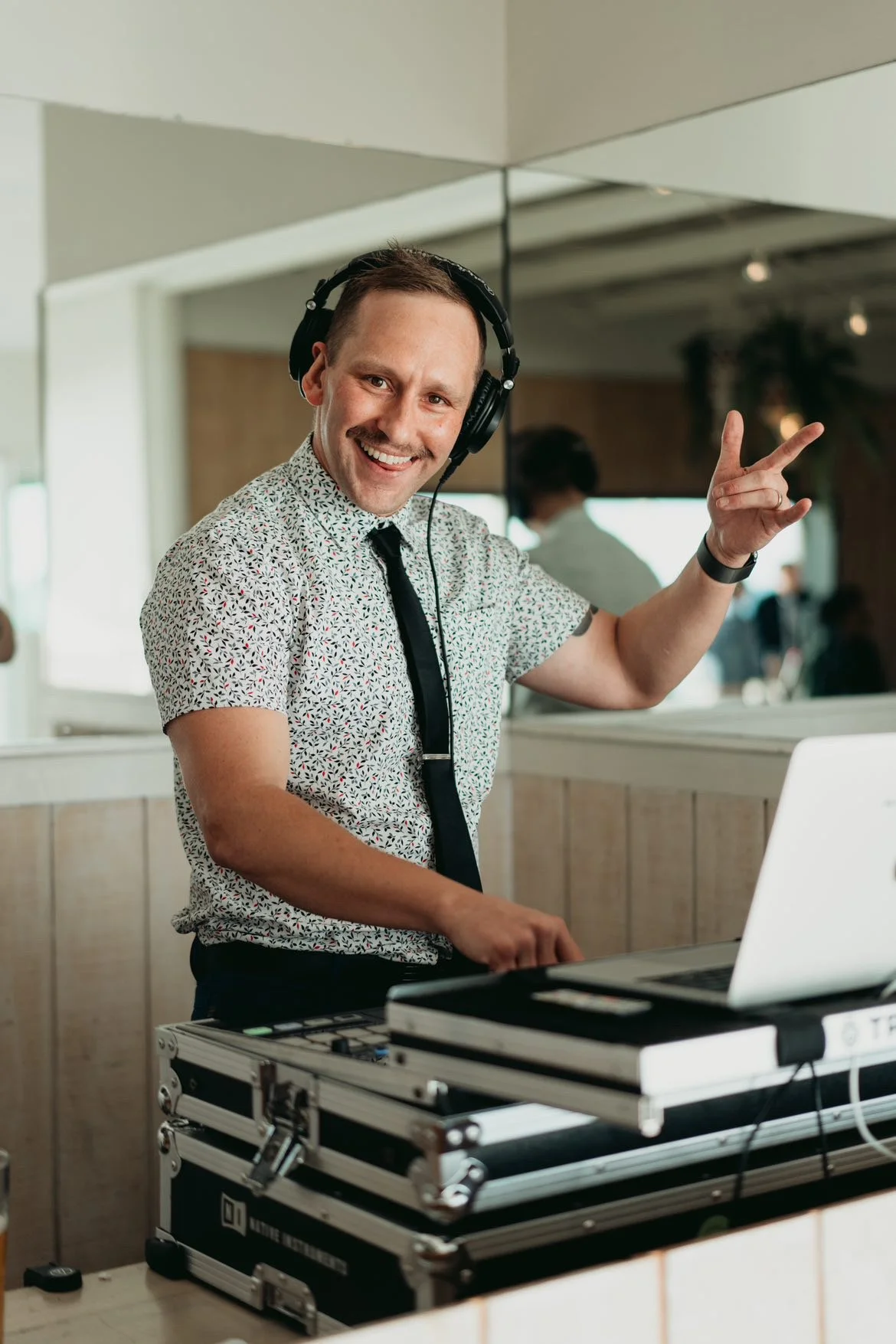 A man smiling and making a peace sign with his right hand while DJing with headphones on in a lively indoor setting.