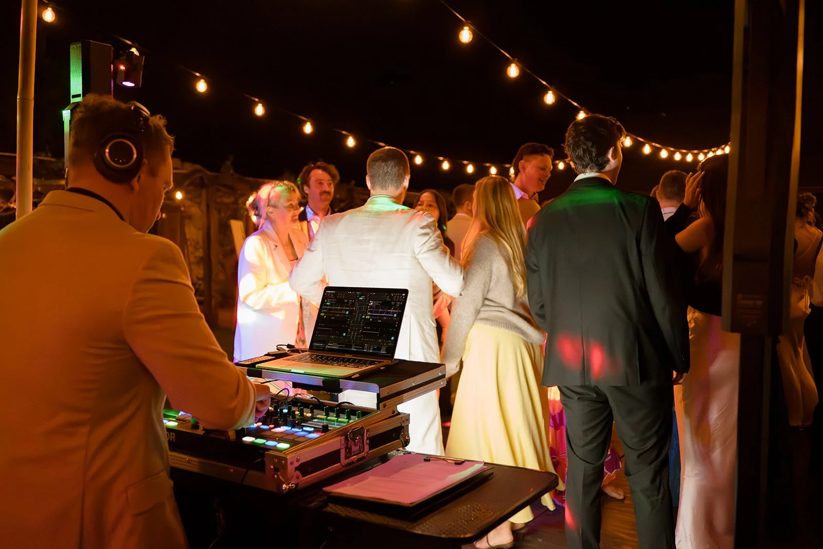 DJ in a yellow jacket playing music at an outdoor party with string lights overhead and people dancing in formal attire at night.