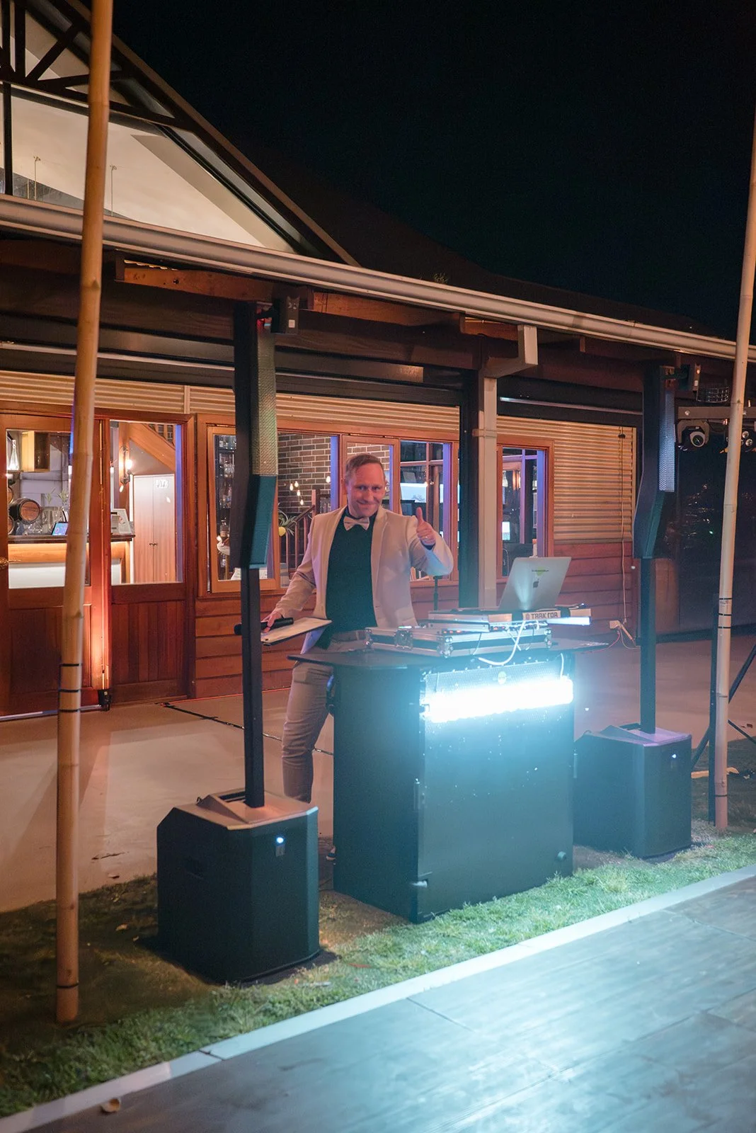 A man in a white jacket giving a thumbs up behind DJ equipment at an outdoor event at night.