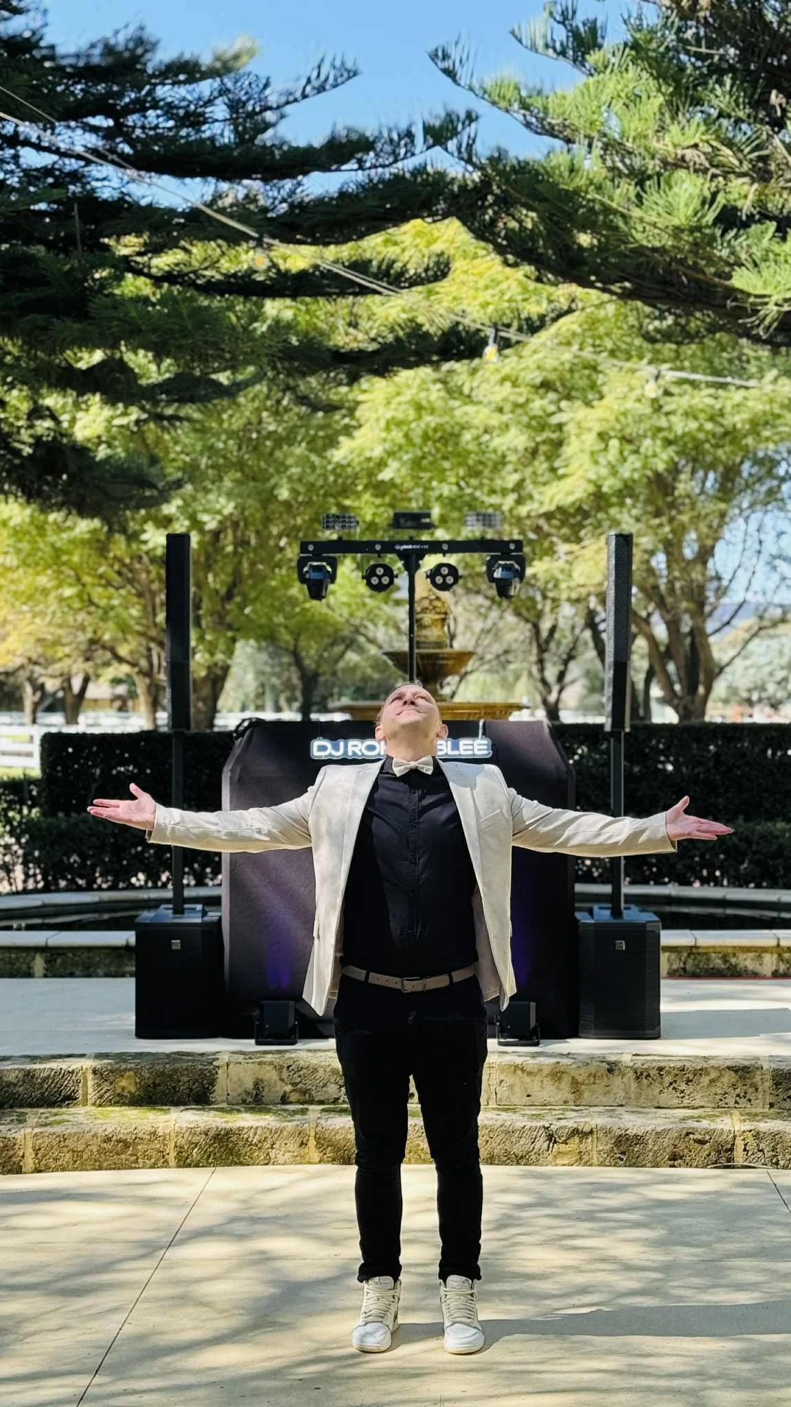 A man wearing a white blazer, black shirt, and beige shoes is standing outdoors in front of a DJ booth with his arms outstretched, enjoying the sunlight and nature.