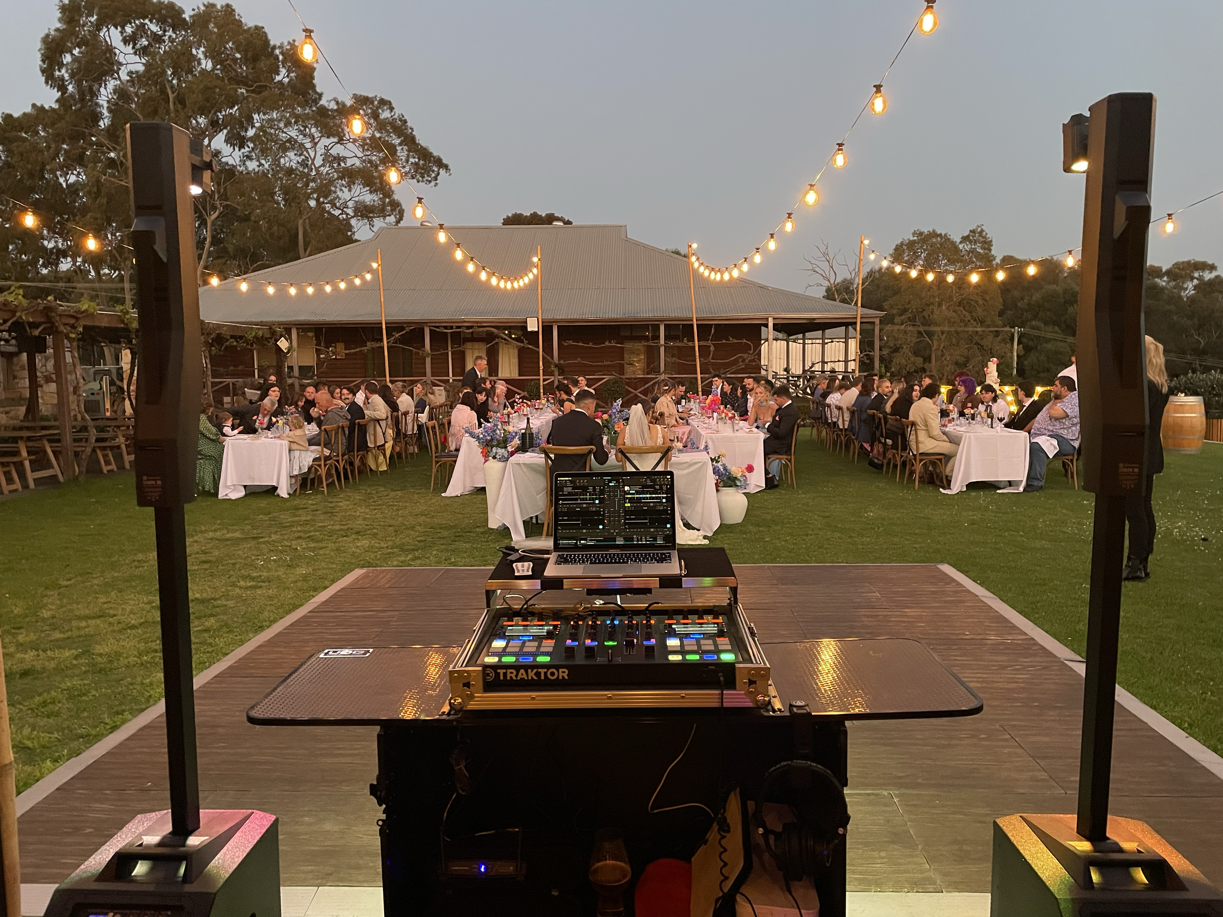 Outdoor evening wedding reception with tables decorated with flowers, string lights overhead, and guests seated, seen from the DJ's viewpoint with equipment in the foreground.