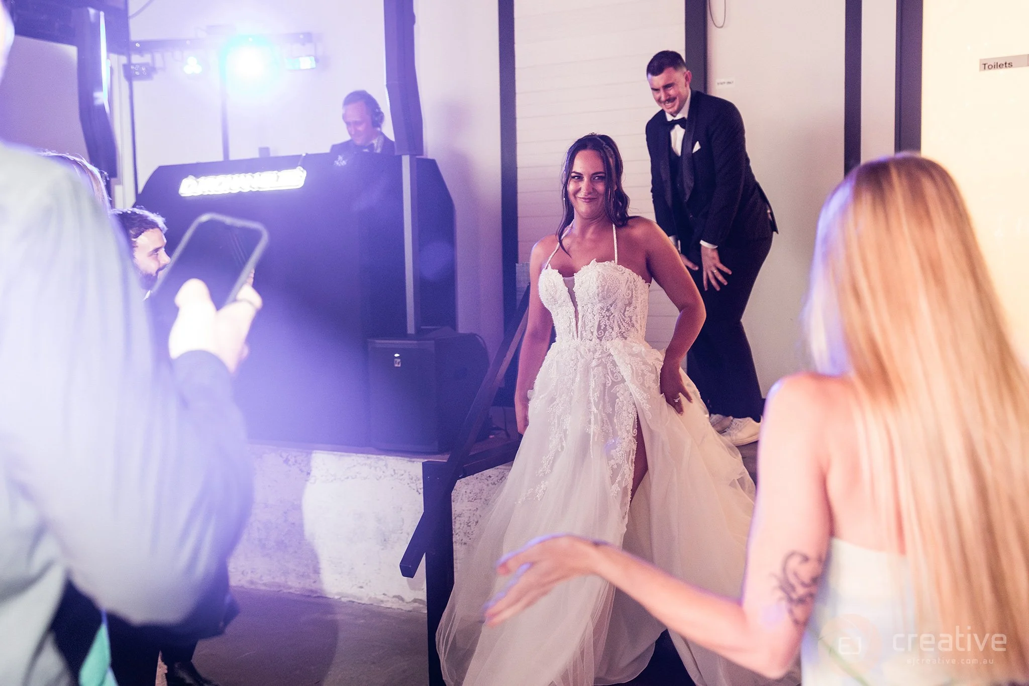 A bride in a white lace wedding gown dancing at her wedding reception, with a groomsman in a black tuxedo smiling behind her. Guests are taking photos and watching, with DJ equipment and a DJ in the background.