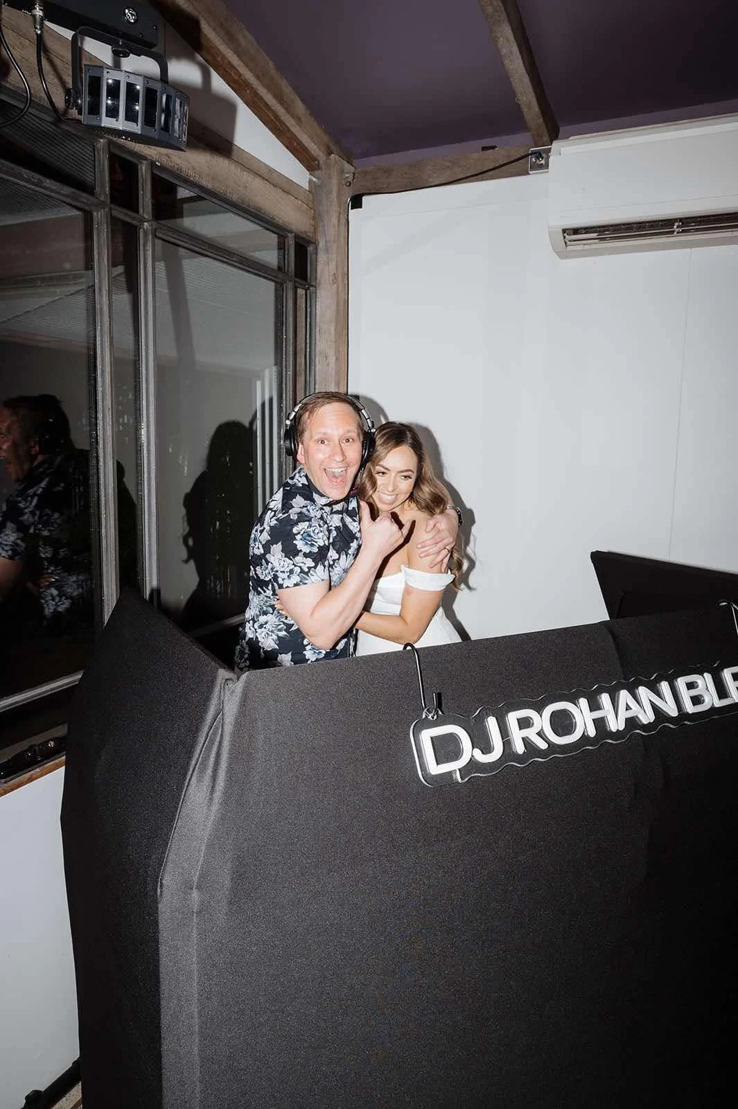 A man and woman smiling and embracing behind a DJ booth with a sign that reads 'DJ Rohan Ble' in a room with white walls, a window, and air conditioning unit.