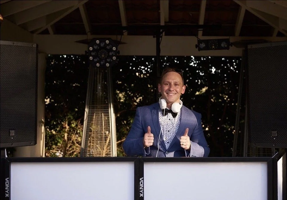 A man in a blue suit with headphones around his neck standing behind a DJ booth, smiling and giving a thumbs-up.