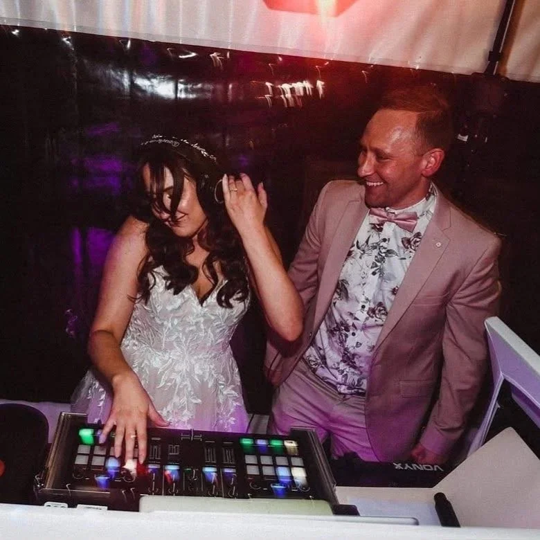 Photo of DJ Rohan Blee standing behind his booth with bride wearing headphones pretending to DJ.