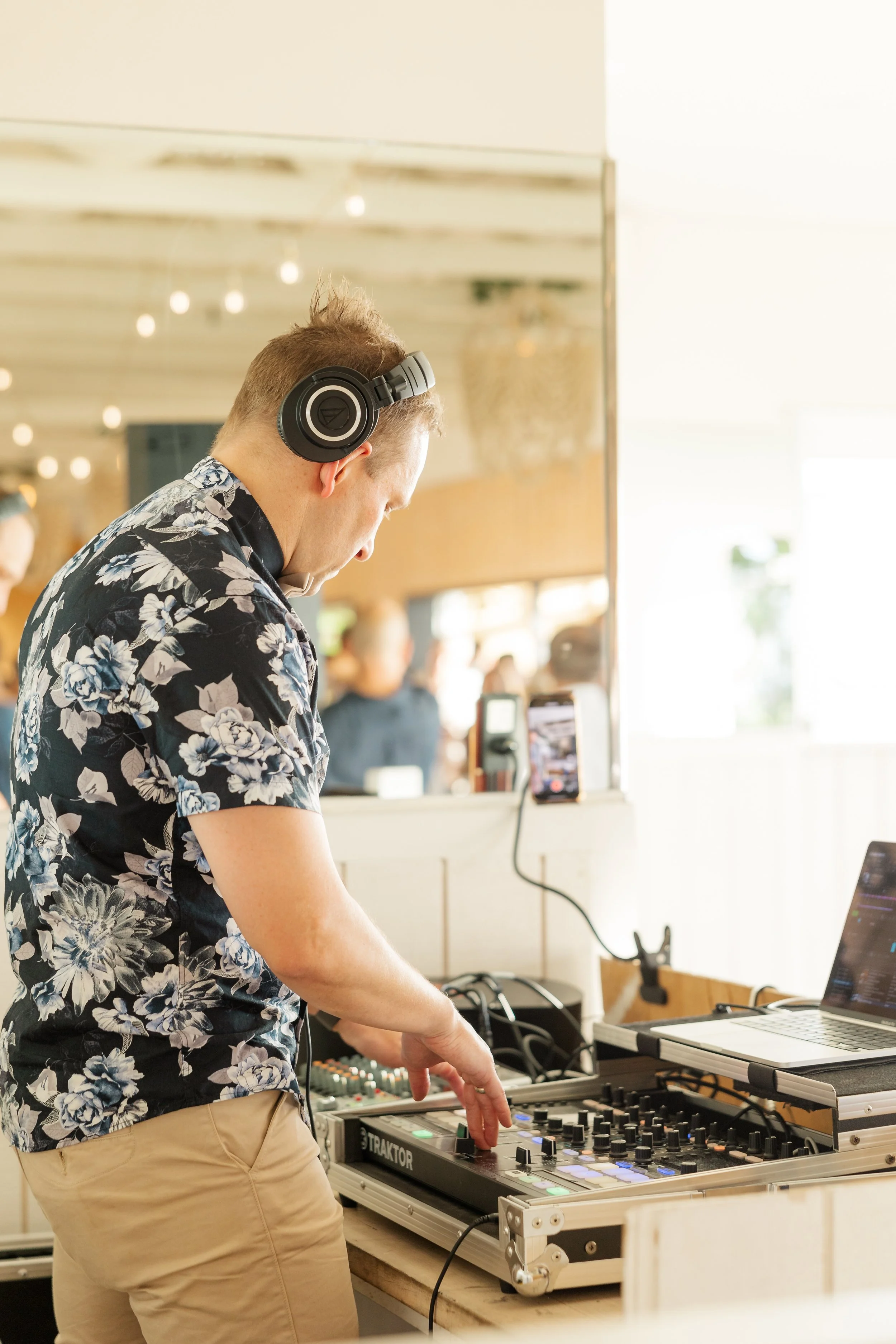 A DJ wearing headphones and a floral shirt mixing music on a DJ controller with a laptop nearby at a lively indoor event.