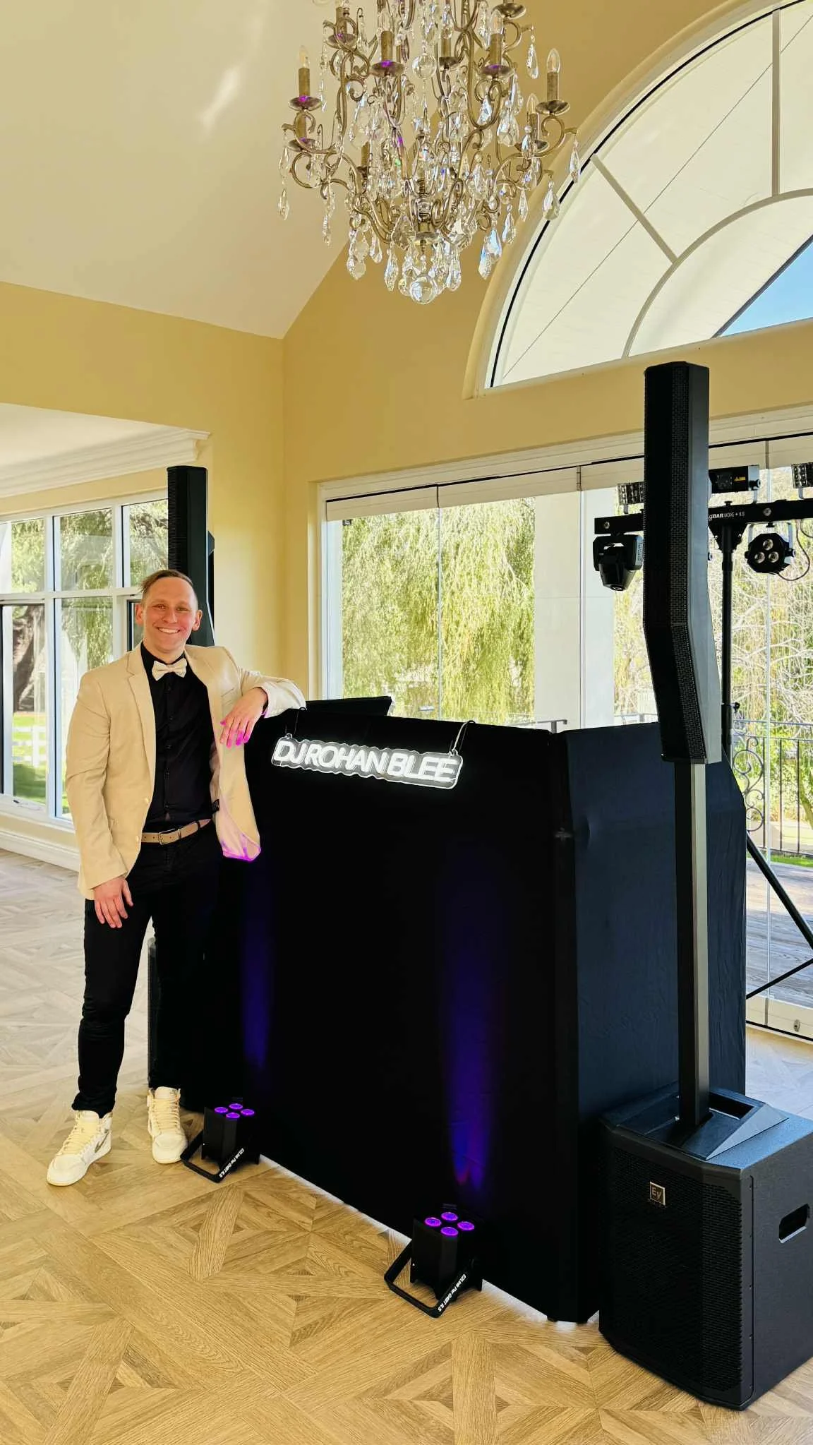 A man in a tuxedo with a bow tie, beige blazer, and white shoes standing next to a DJ booth with a sign that reads 'Durohanblee.' The man is smiling and posing indoors near large windows with a chandelier hanging from the ceiling.