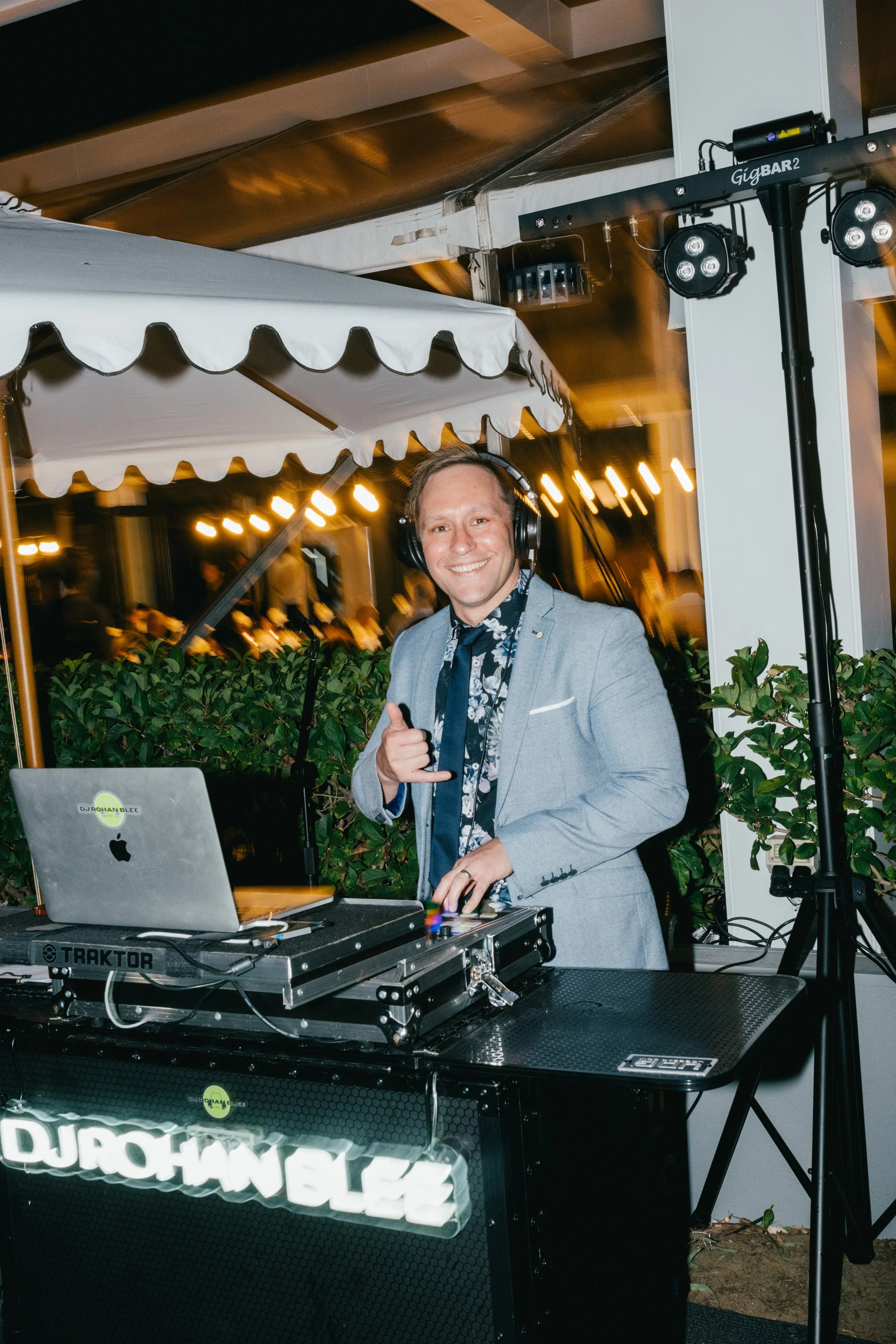 A smiling man in a light gray suit and floral shirt DJing at an outdoor event, with DJ equipment and a laptop on the table, illuminated by string lights at night.