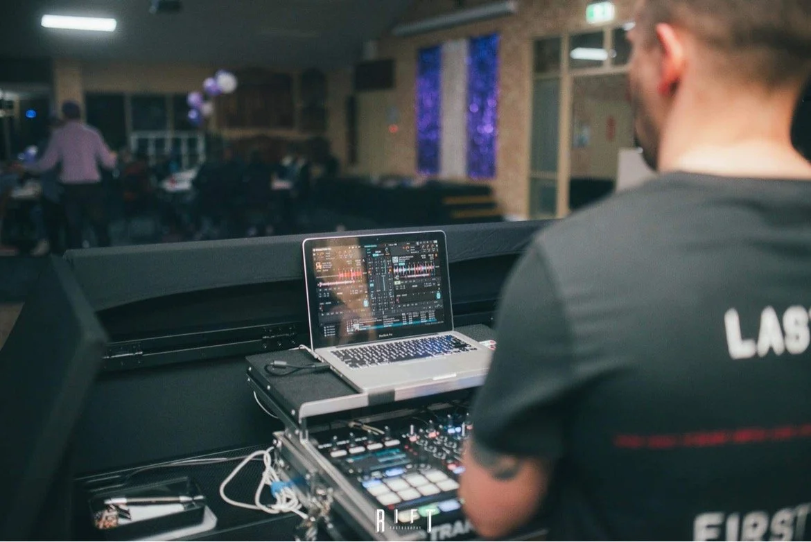 DJ using a laptop and mixer at a nightclub or event, with guests in the background.