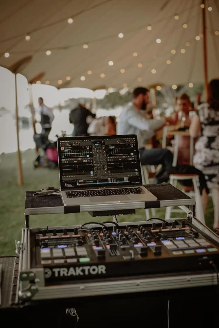 DJ equipment and a laptop set up in front of a blurred outdoor party scene with people sitting under a tent with string lights.