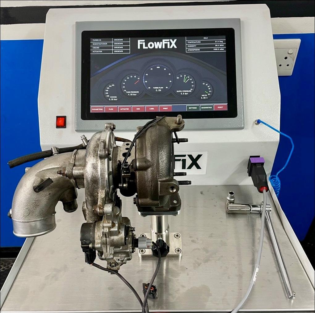 A test bench with a turbocharger connected to a FlowFiX monitoring system. The system's digital screen displays gauges and data related to turbo performance, including pressure, temperature, humidity, and voltage.