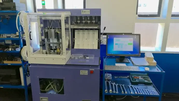 Laboratory or industrial equipment setup with a machine connected to a computer on a blue desk, with tools and components laid out nearby.