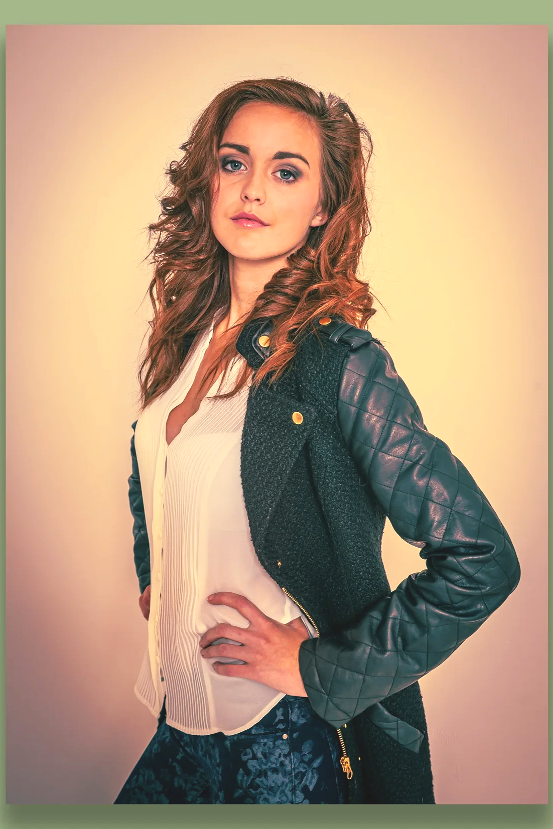A young woman with wavy brown hair and blue eyes posing with her hand on her hip, wearing a black leather jacket with quilted sleeves over a white blouse, standing against a gradient background.