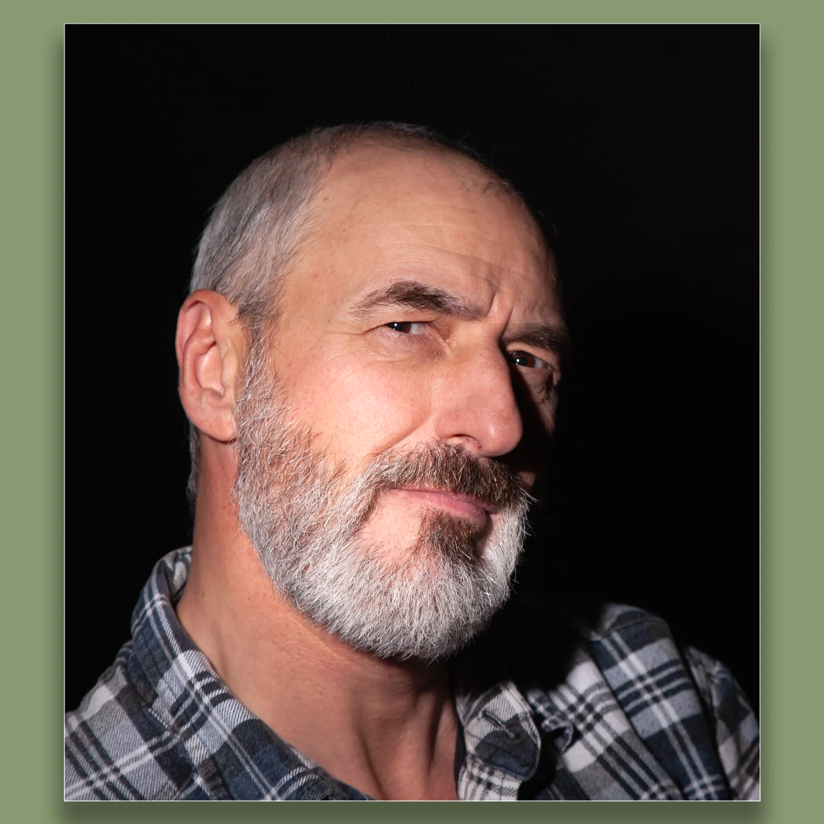 Close-up portrait of a middle-aged man with a gray beard and mustache, wearing a plaid shirt, looking confidently at the camera against a black background.