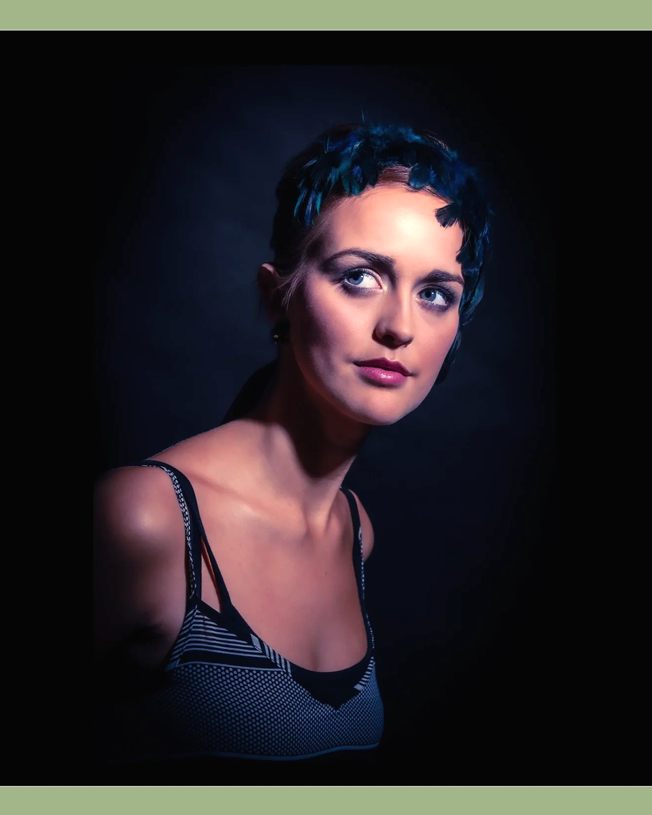 A woman with blue highlights in her short, dark hair, wearing a striped sleeveless top, looking to her right against a dark background.