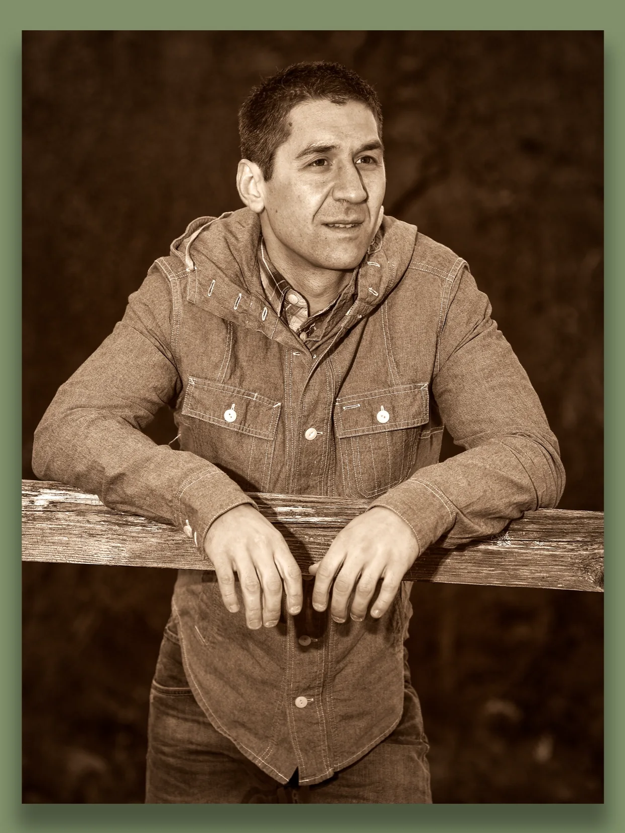 A man in a denim jacket and plaid shirt leaning on a wooden fence outdoors, looking to the side with a thoughtful expression, sepia tone photo.